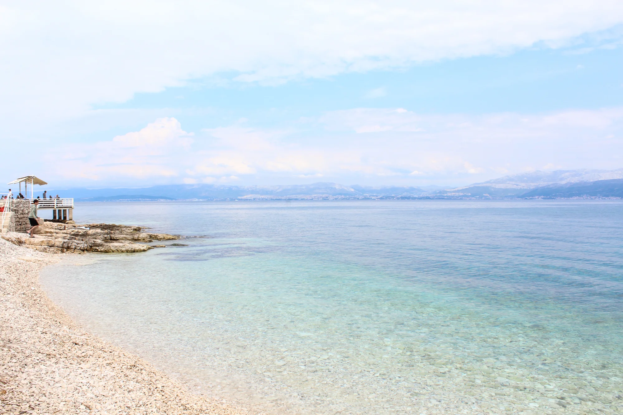A peaceful beach with clear, shallow water and a rocky shoreline. In the background, a pier with umbrellas and people enjoying the view. Distant mountains are visible under a partly cloudy sky.