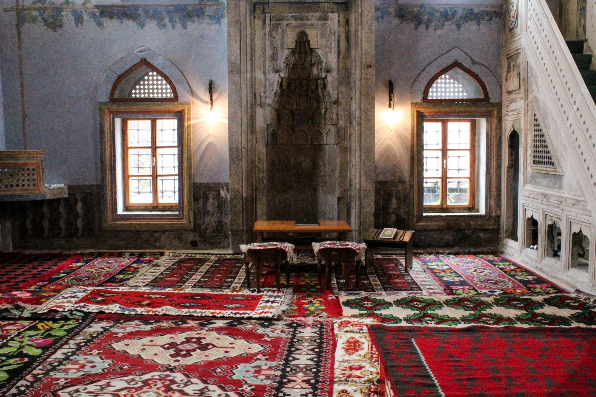 Interior of a room decorated with various colorful patterned rugs on the floor, with two windows, a small table, and two chairs. The walls are aged with decorative trim and a central niche.