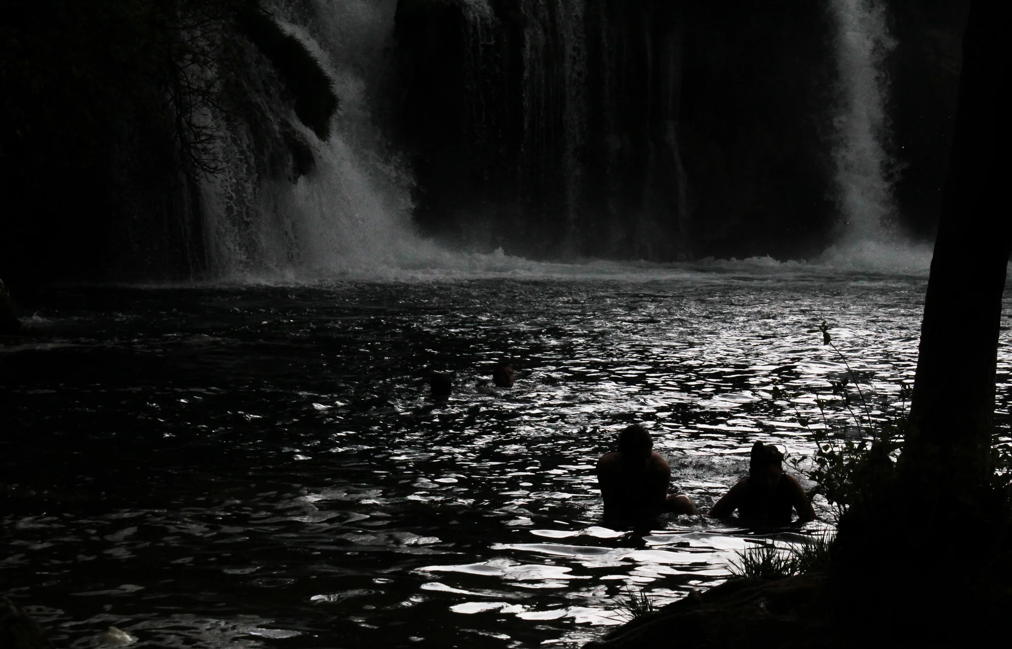 People swimming near a waterfall in a dark, shaded environment.