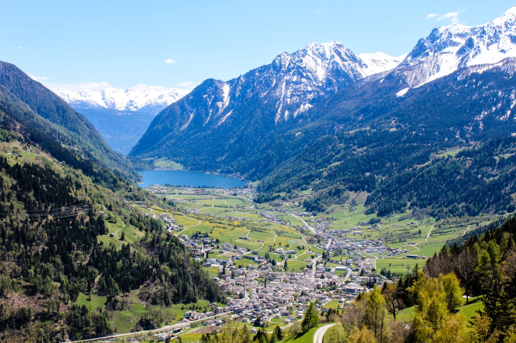 A scenic view of a mountain valley with snow-capped peaks, a lake, and a small town with green fields and trees.
