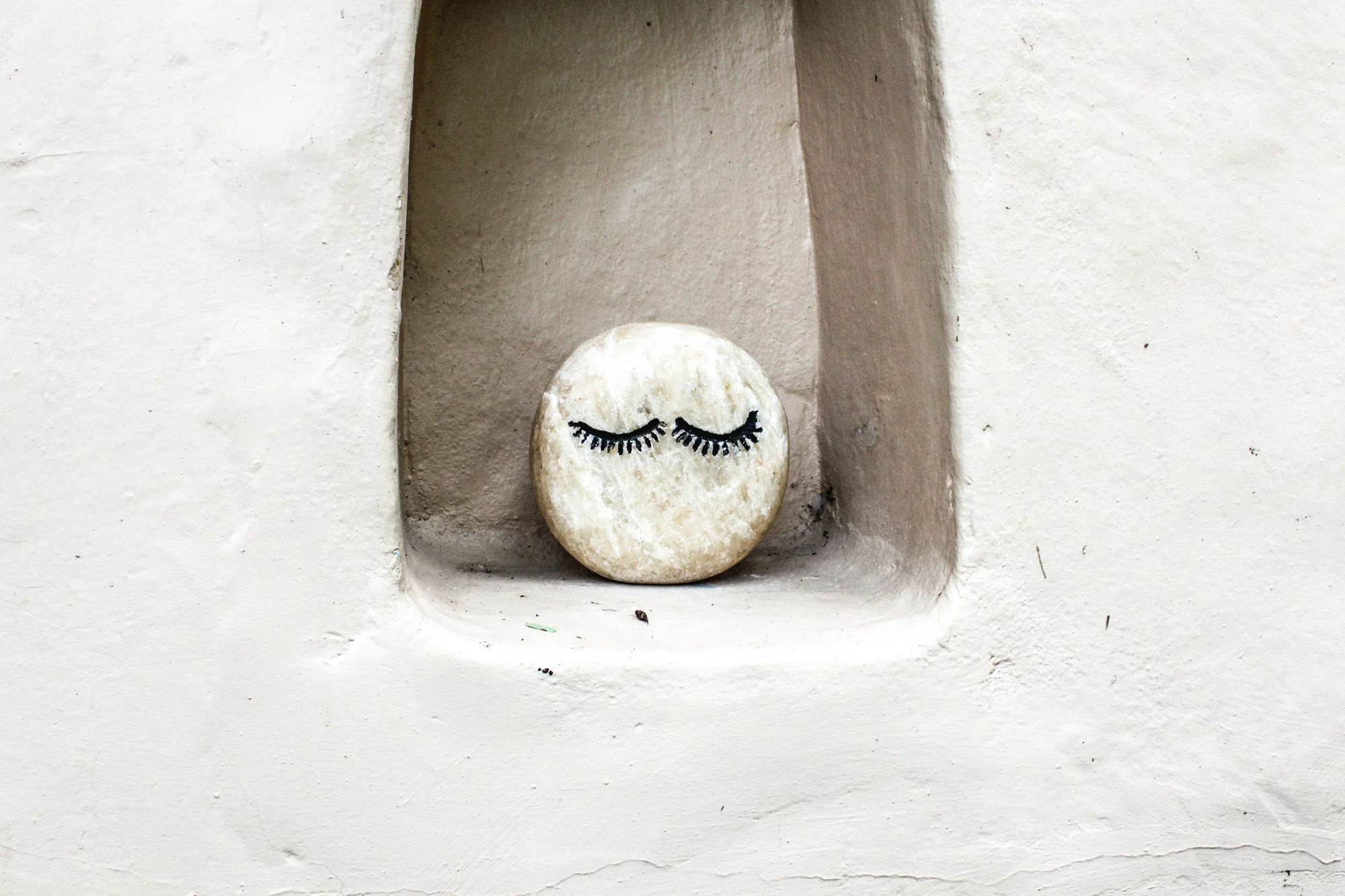 A small, round stone with a painted smiling face and closed eyes with long eyelashes. The stone is placed in a wall nook.