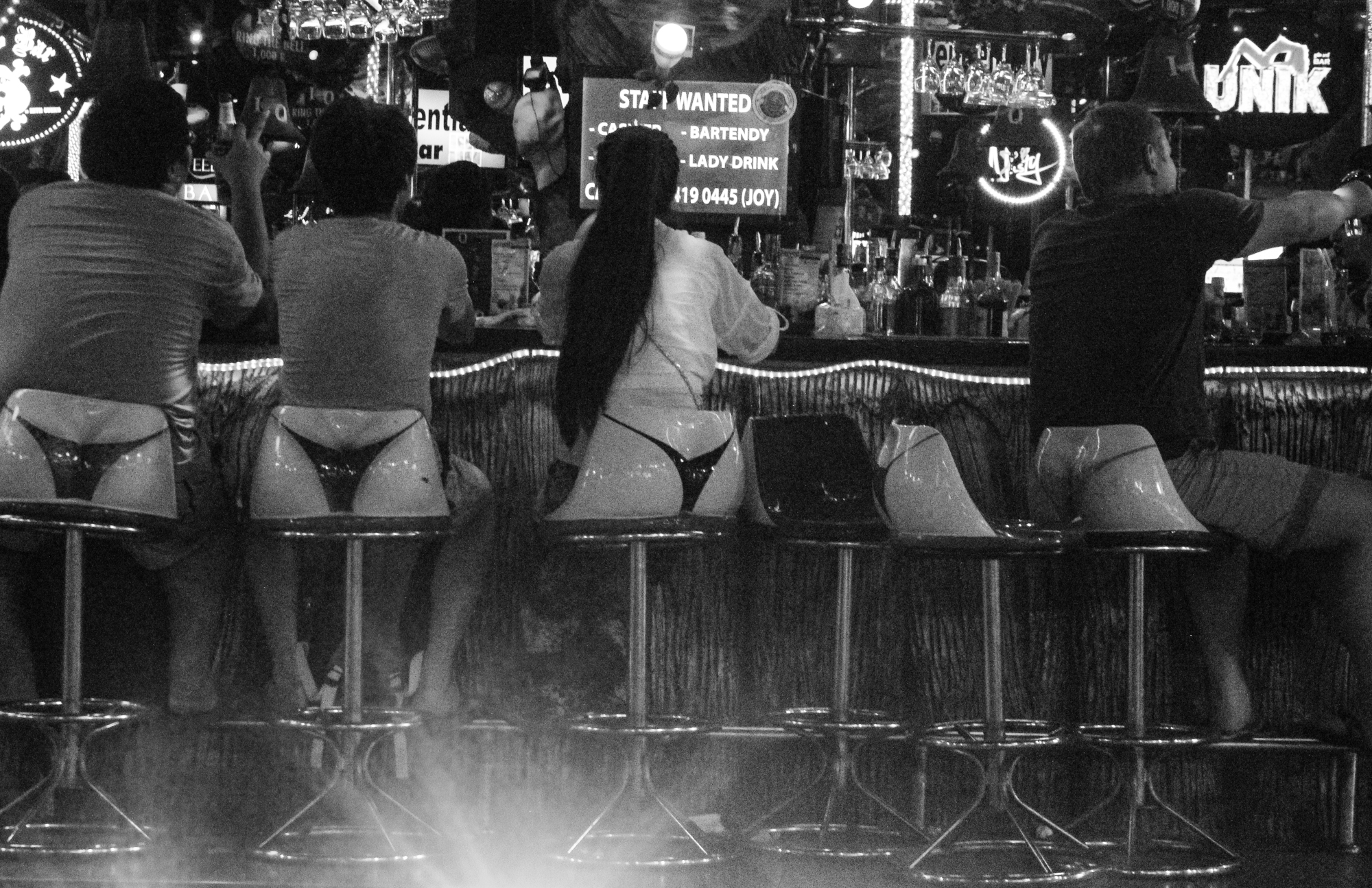 Bar scene with four people sitting at the counter, two women wearing revealing outfits, and a man wearing a T-shirt and shorts. The background has illuminated signs and bottles behind the bar.