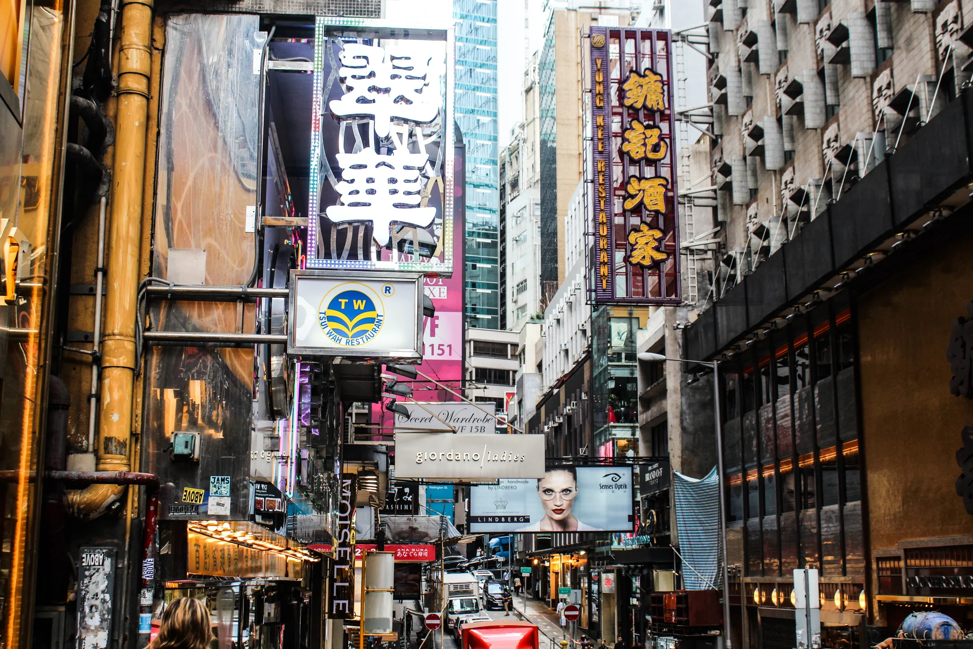 A busy urban street scene with numerous neon signs, billboards, and storefronts in a dense city area, likely in Asia.