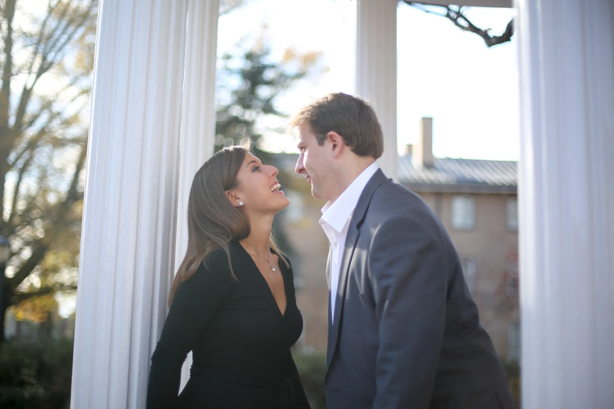 A man and a woman face each other and smile between white columns outside during daytime.