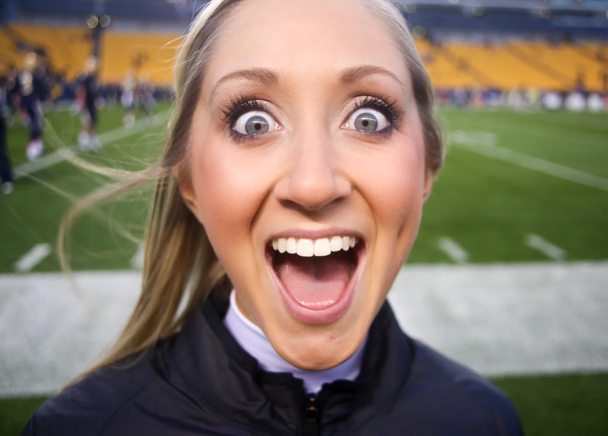 Close-up of a woman with a surprised expression at a football stadium, with blurred football field and players in the background.
