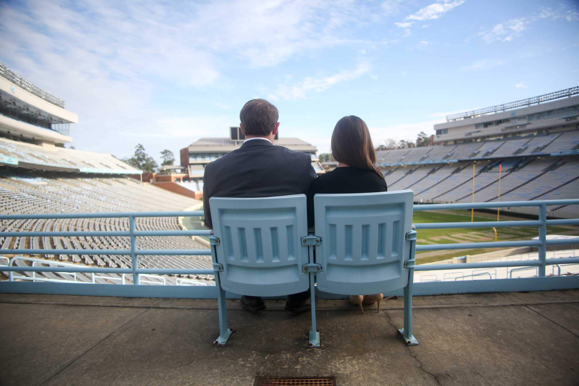 A man and woman sitting on a stadium bench, looking out at an empty football field under a partly cloudy sky.