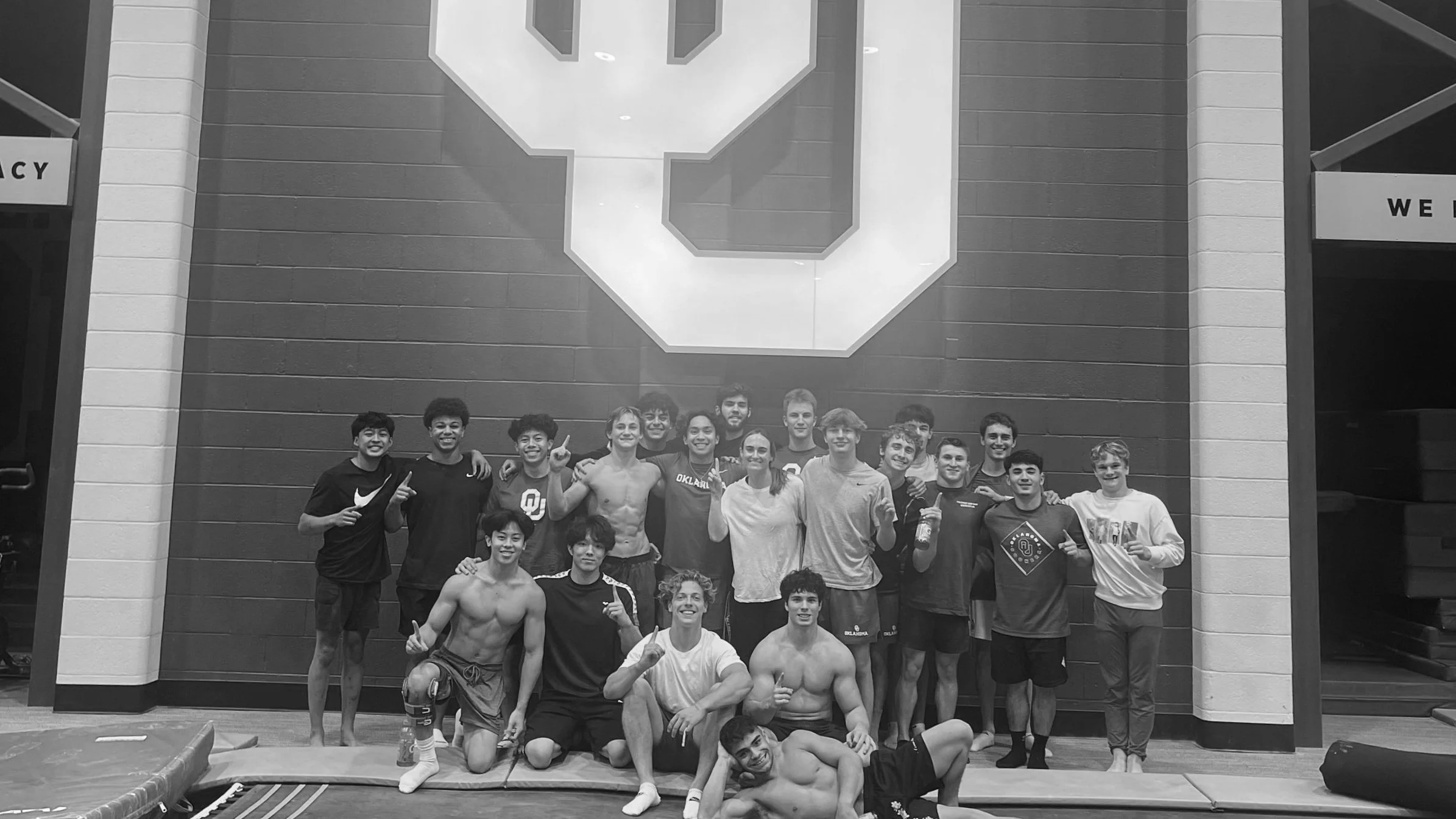 OU men's gymnastics team posing for a photo in an indoor gym facility, some shirtless, in front of a wall with a large University of Oklahoma logo.