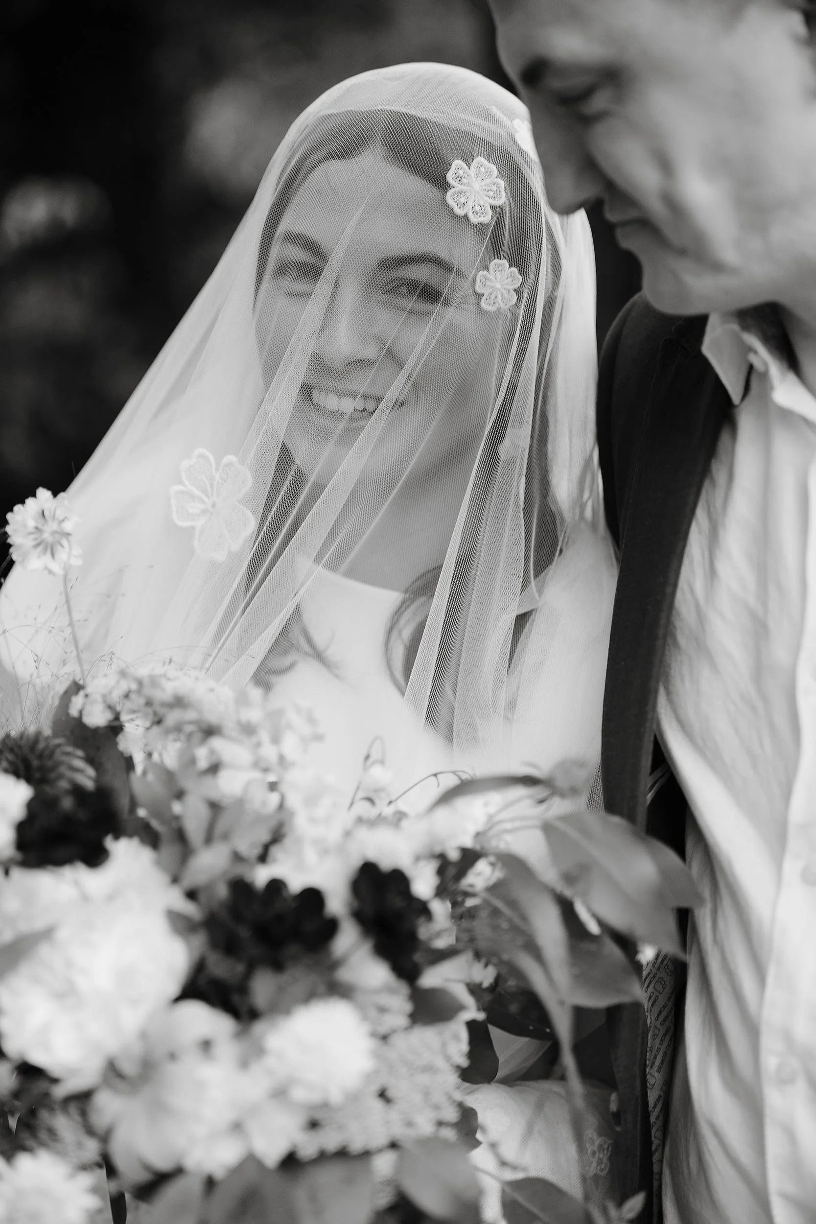 The bride looks out from her designer wedding veil and has a huge beaming smile with her wedding bouquet clutched closely to her heart. The groom dressed in an open neck shirt and jacket has a look of quiet satisfaction.