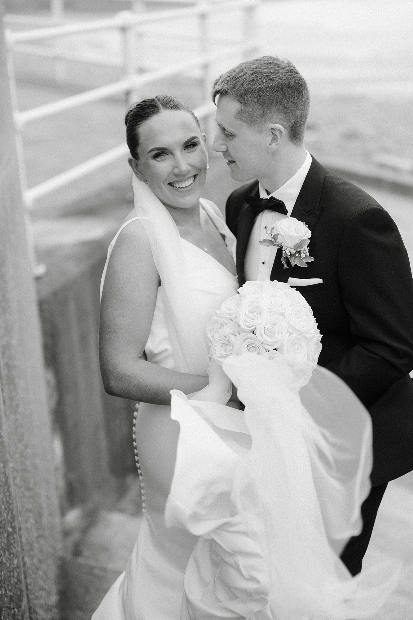 the bride and groom in black and white on the steps down to the beach sheltering from the wind and rain. The bride looks into the camera while holding her bouquet and flowers. The groom looks lovingly into her eyes.