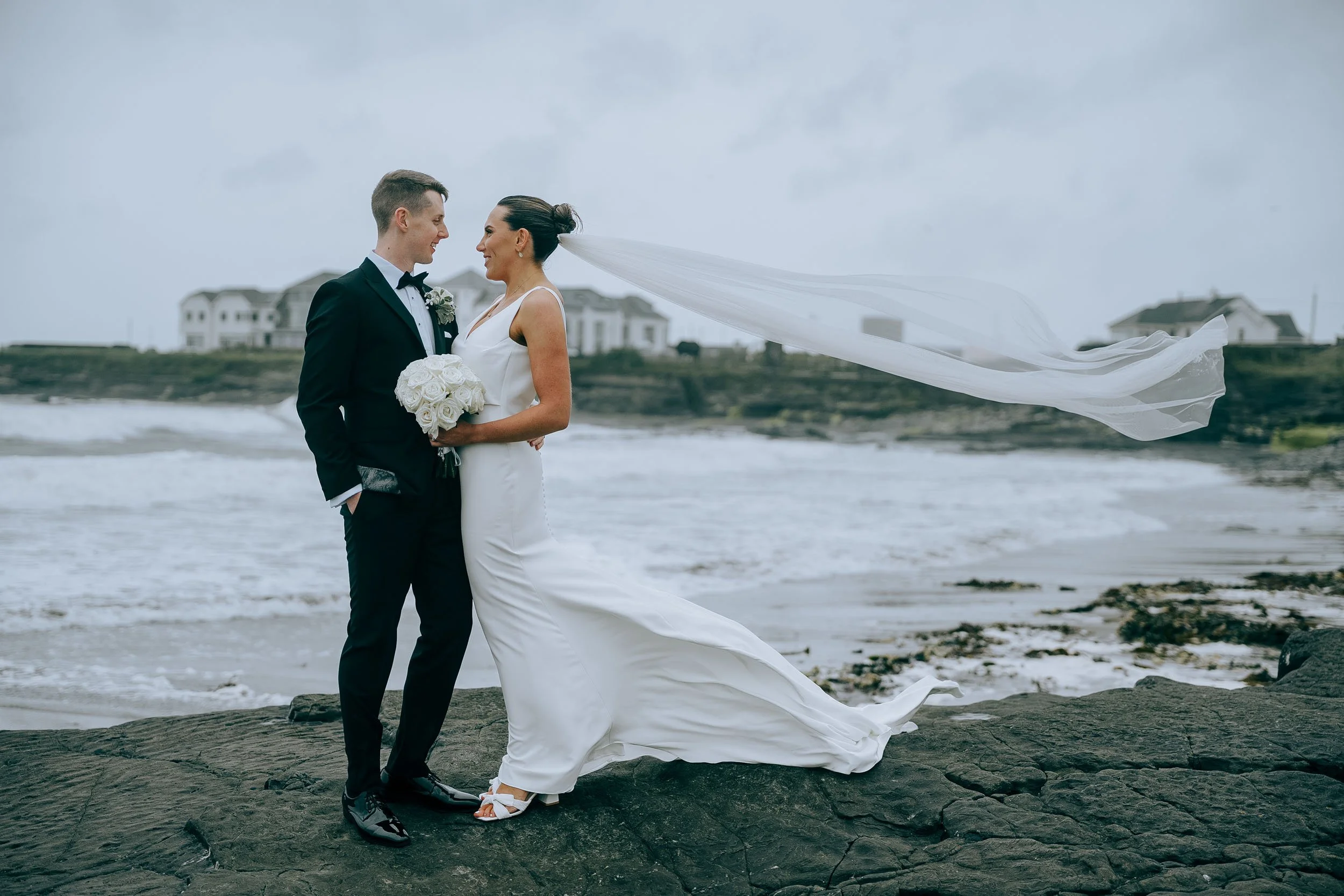 The groom looking very smart in his black tie looking at his bride on the beach with The Armada hotel behind them at Spanish Point.