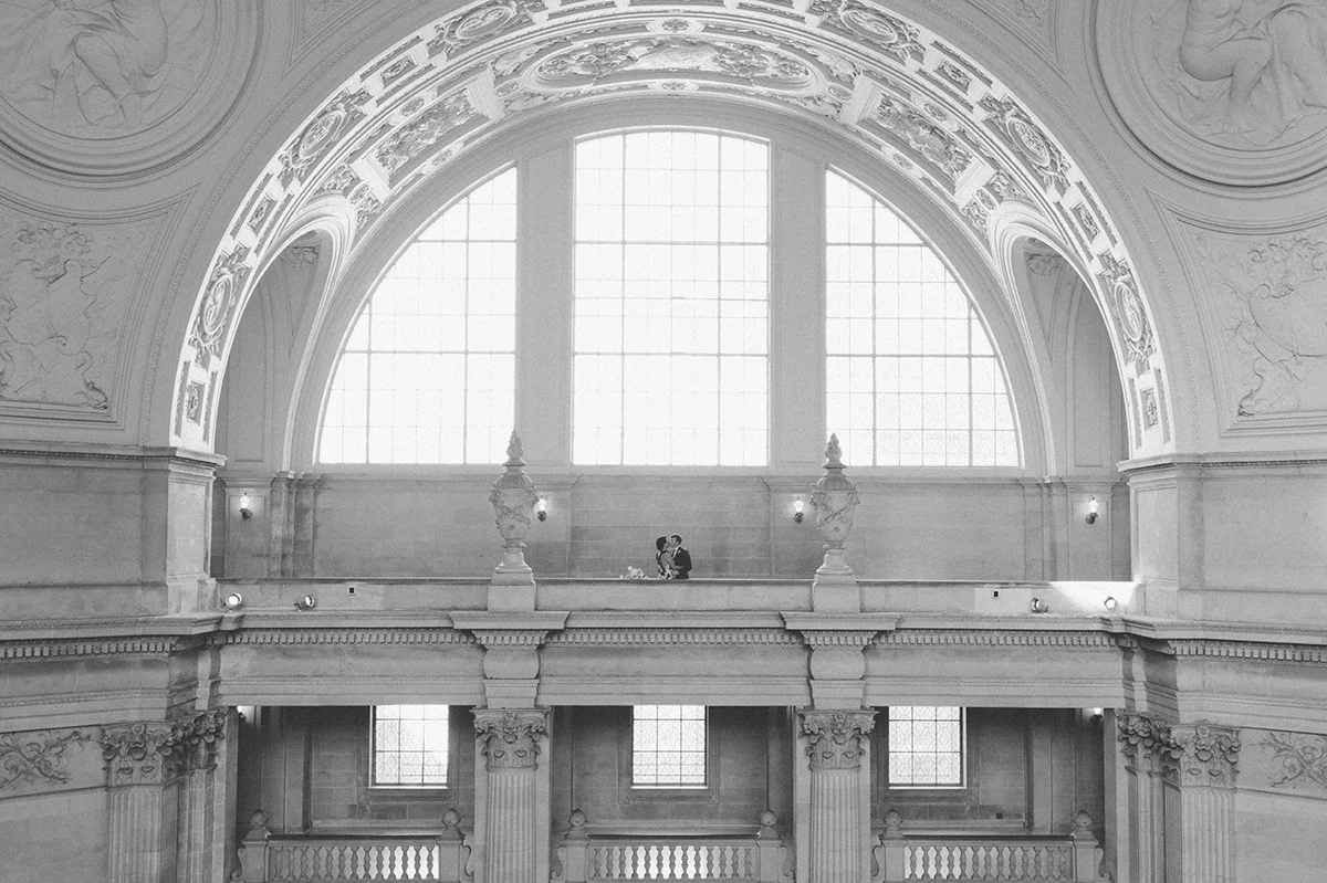 The bride and groom kiss on the 4th floor balcony, under the dramatic arch of San Francisco City Hall.