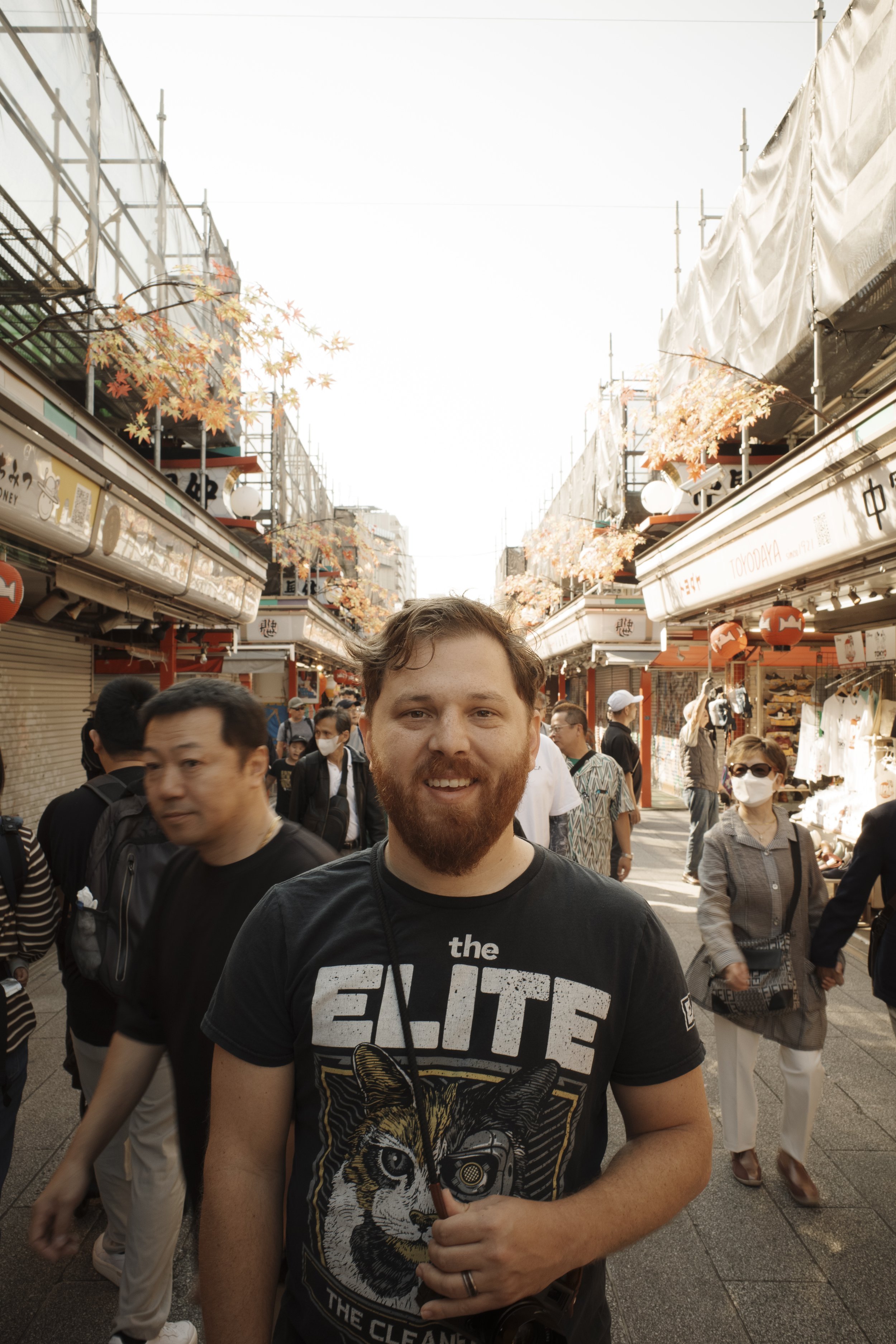 A man with a beard and a camera around his neck smiling at the camera in a busy outdoor shopping street with many people, some wearing masks, and storefronts on either side.
