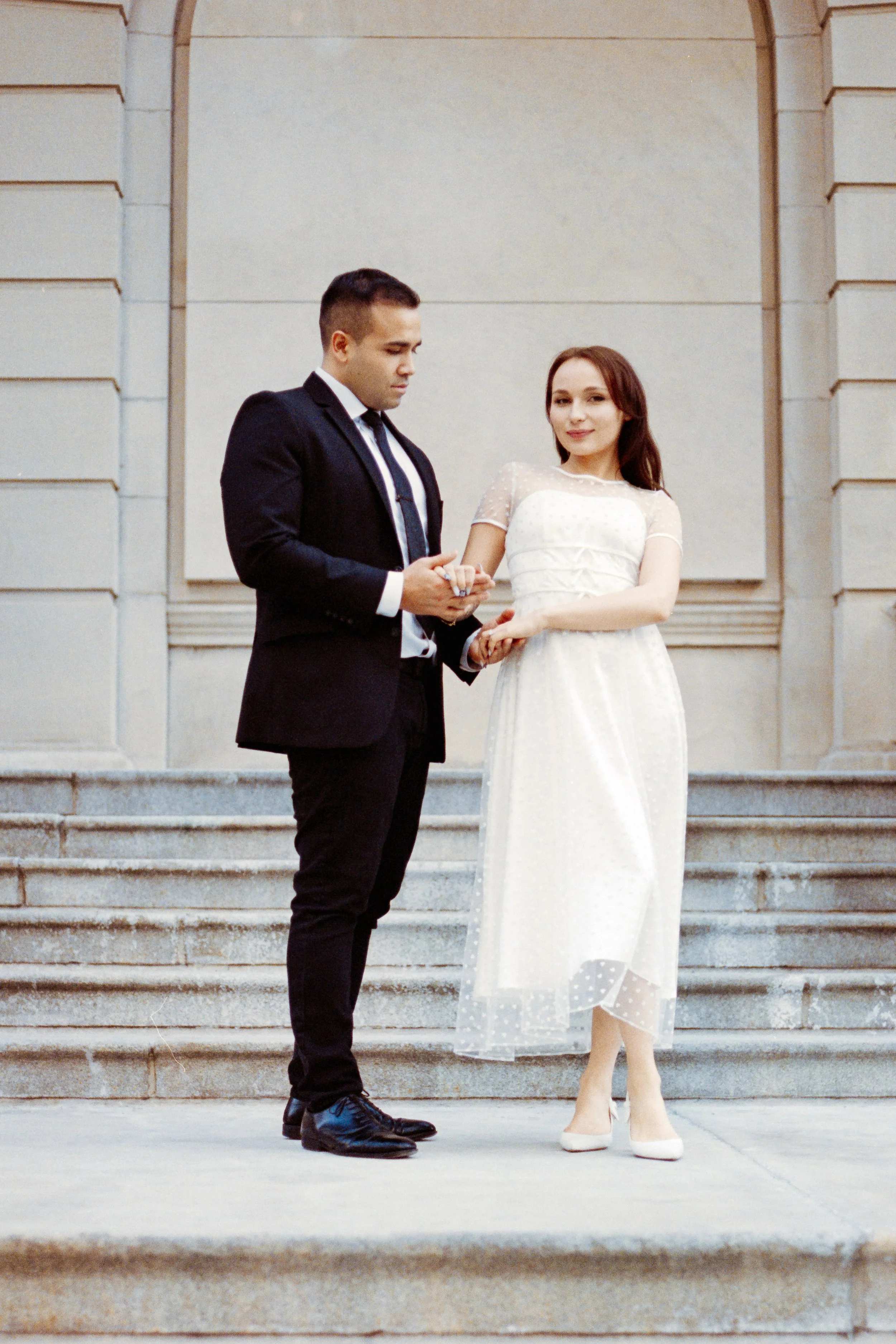 A couple dressed in formal attire standing on building steps; the man in a black suit and the woman in a white dress, during a wedding ceremony.