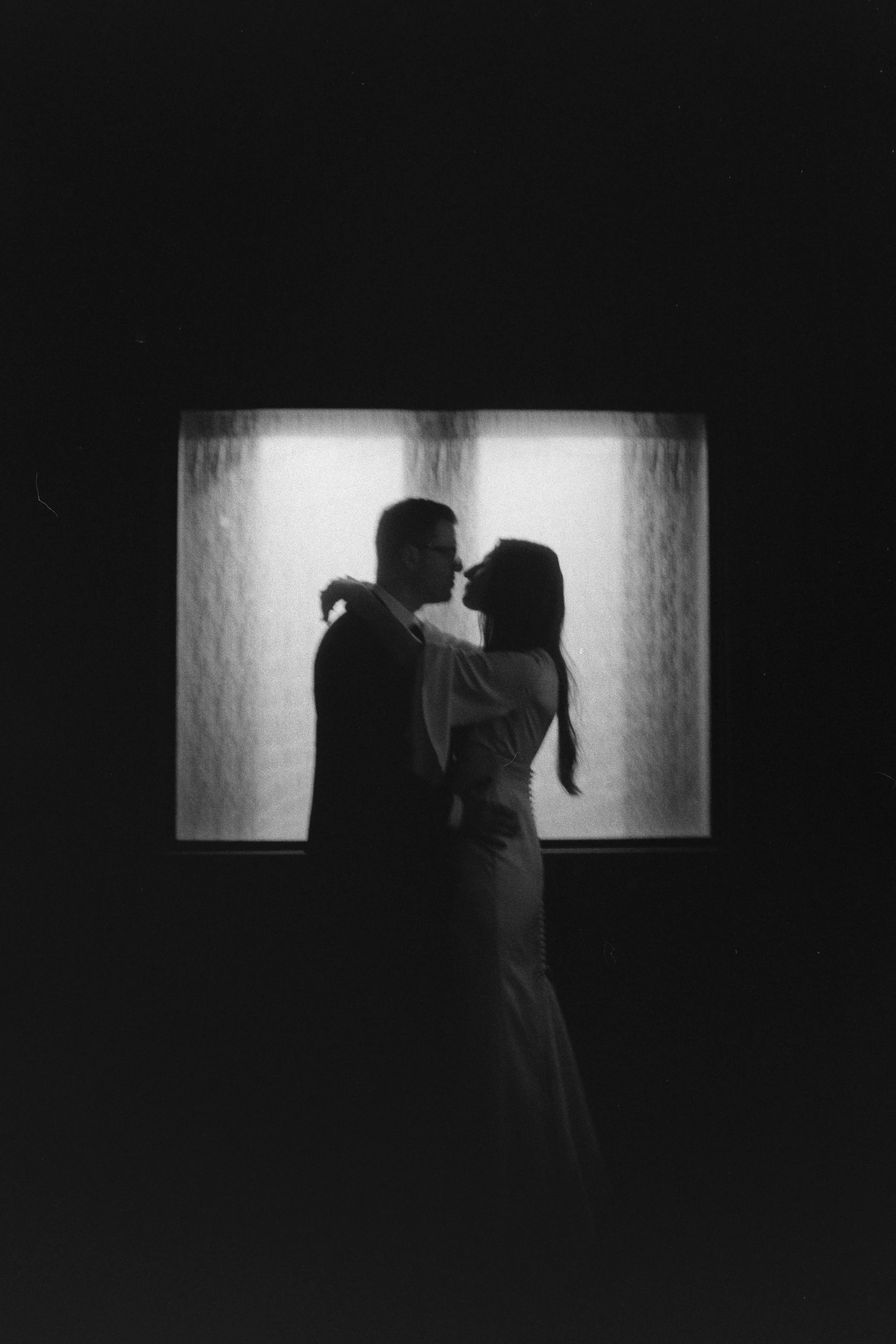 Silhouette of a couple embracing and about to kiss in front of a backlit square window or screen.