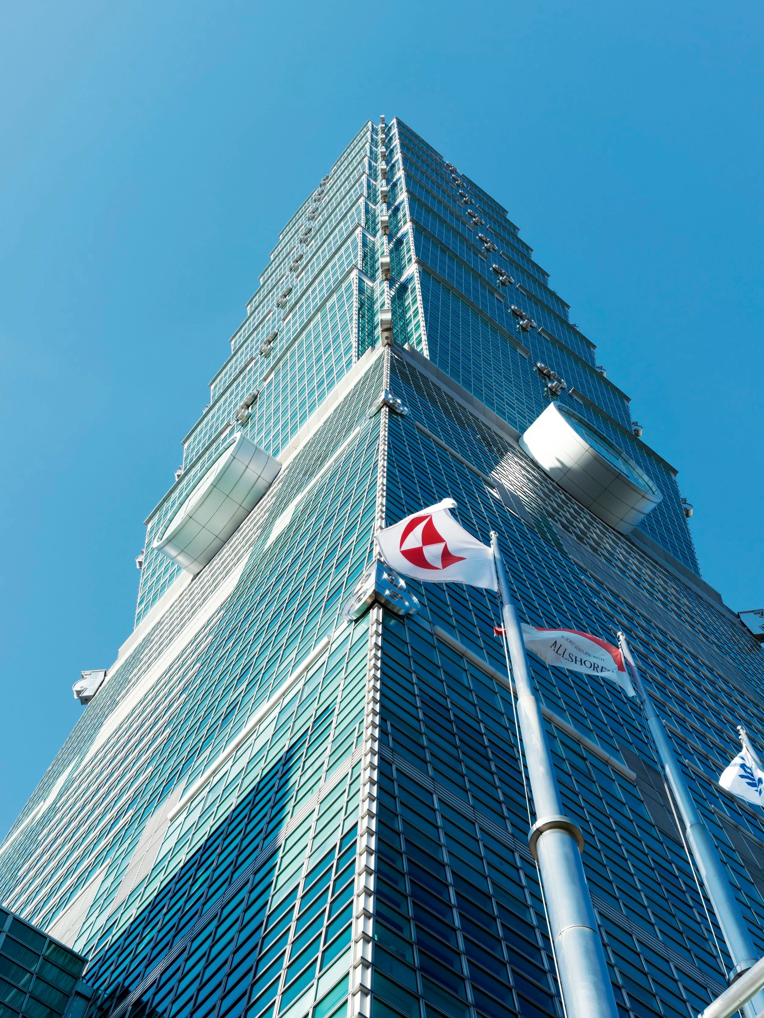 Taipei 101 skyscraper viewed from below against a bright blue sky with flags in front
