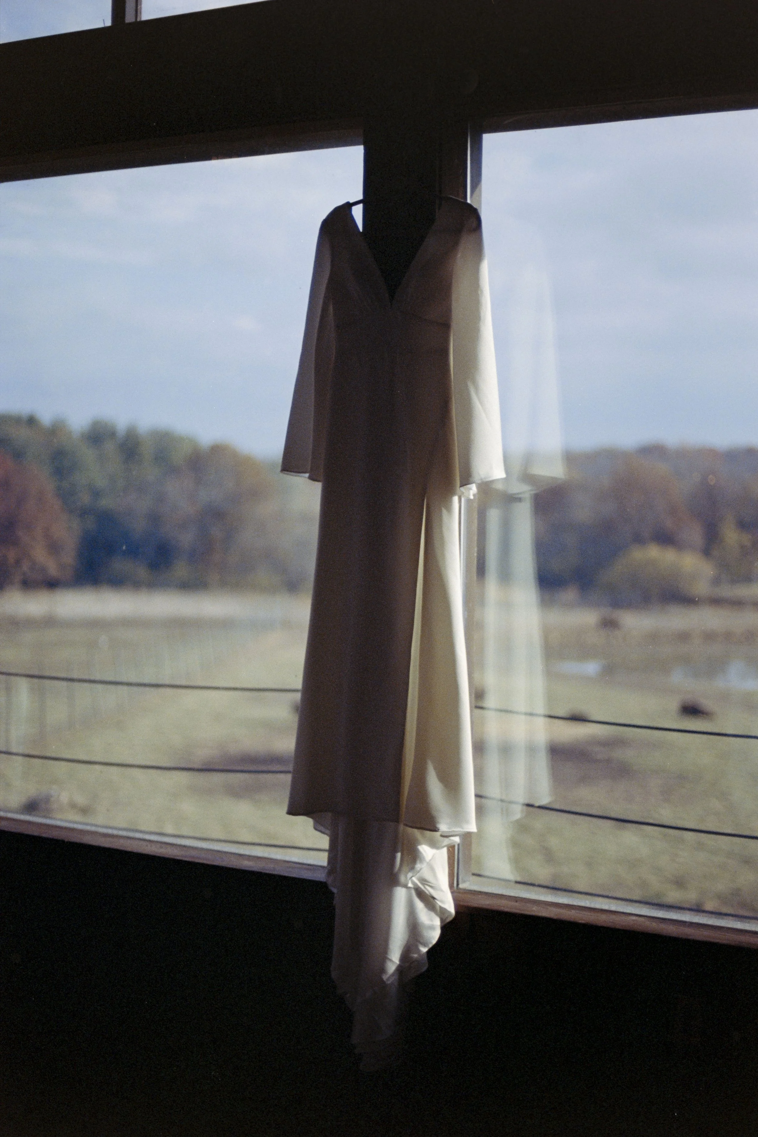 A beige and white garment hangs on a hook in front of a large window with a view of a rural landscape with trees and open fields.