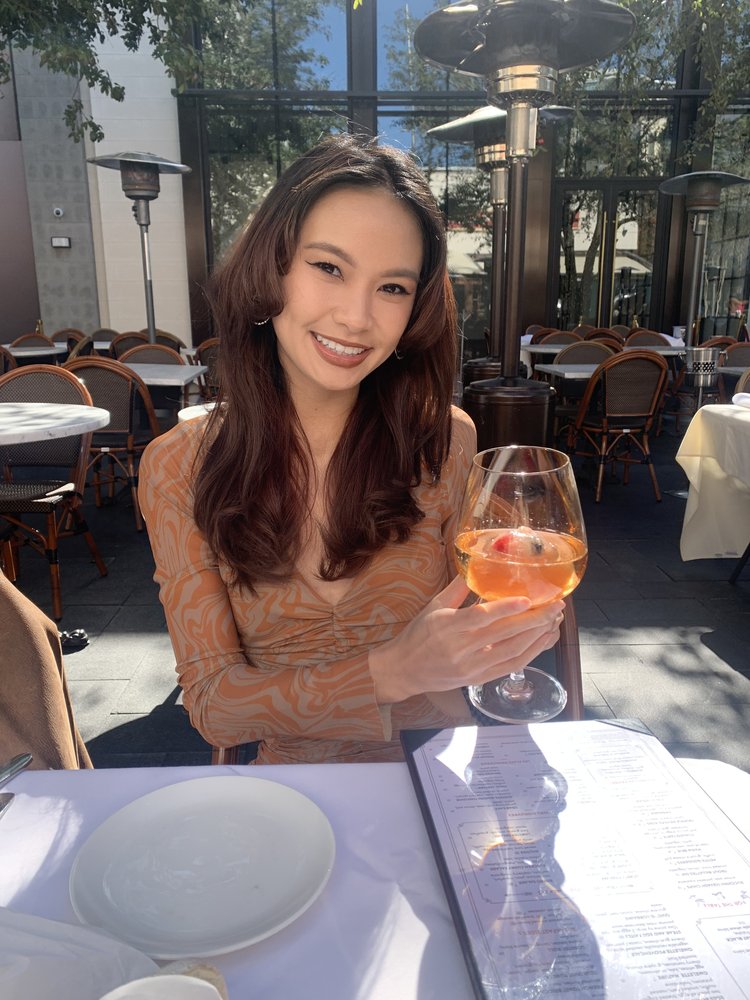 Jocelyn, smiling, sitting at a cafe table under the sun with a glass of wine