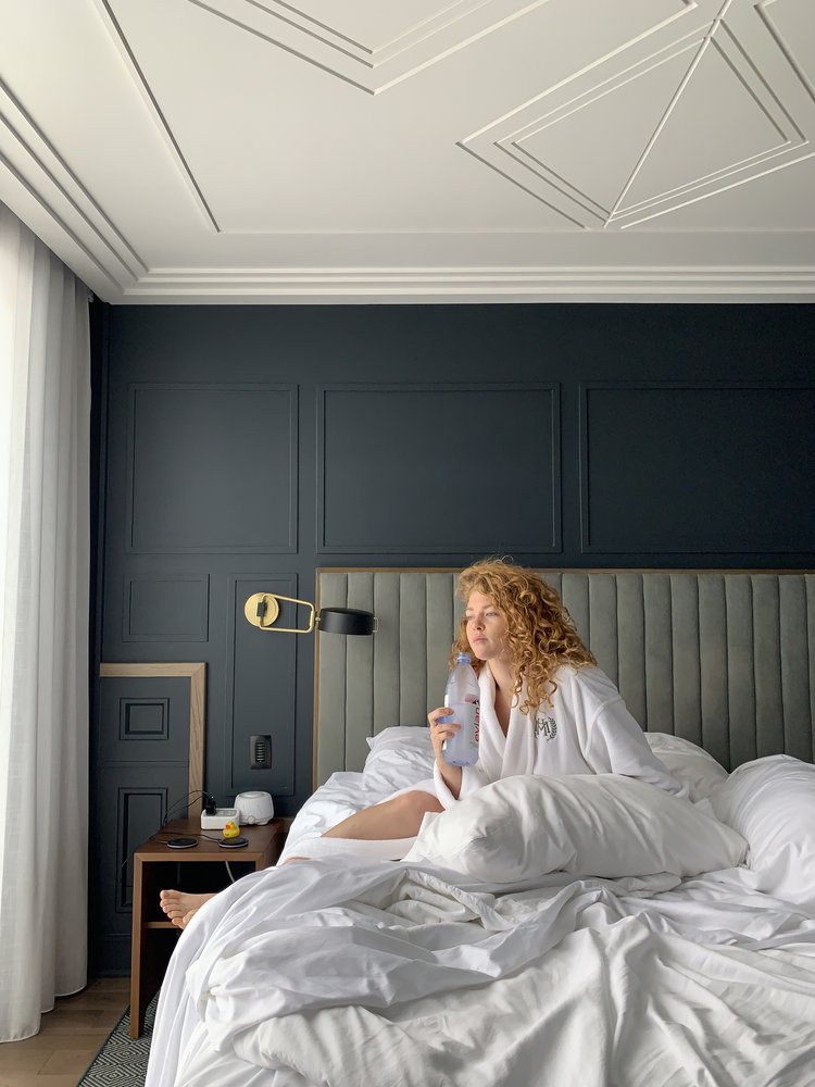 Founder and CEO, Anna Trammell, sitting a bed with a white comforter and in a white robe, looking out the window holding a bottle of water.