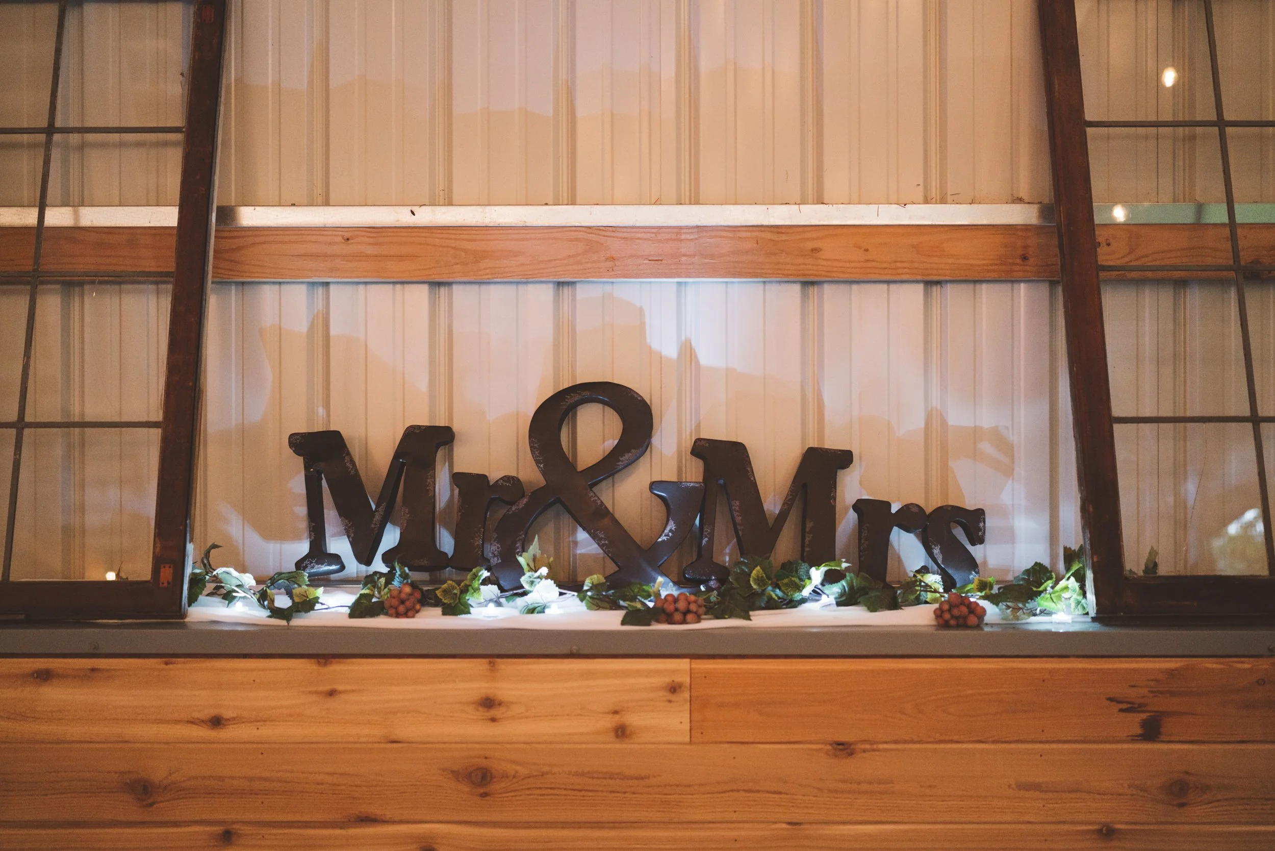 Decorative sign with the words "Mr & Mrs" on a shelf with green leaves and small clusters of red berries, in front of a window with wooden framing.