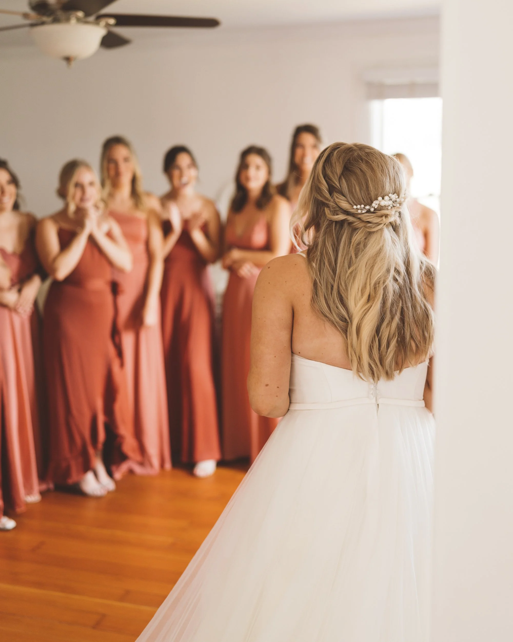 Bride in a white wedding dress with wavy blonde hair and a floral hairpiece, facing a group of bridesmaids in matching reddish-brown dresses, in a bright room with wooden floors and a ceiling fan.