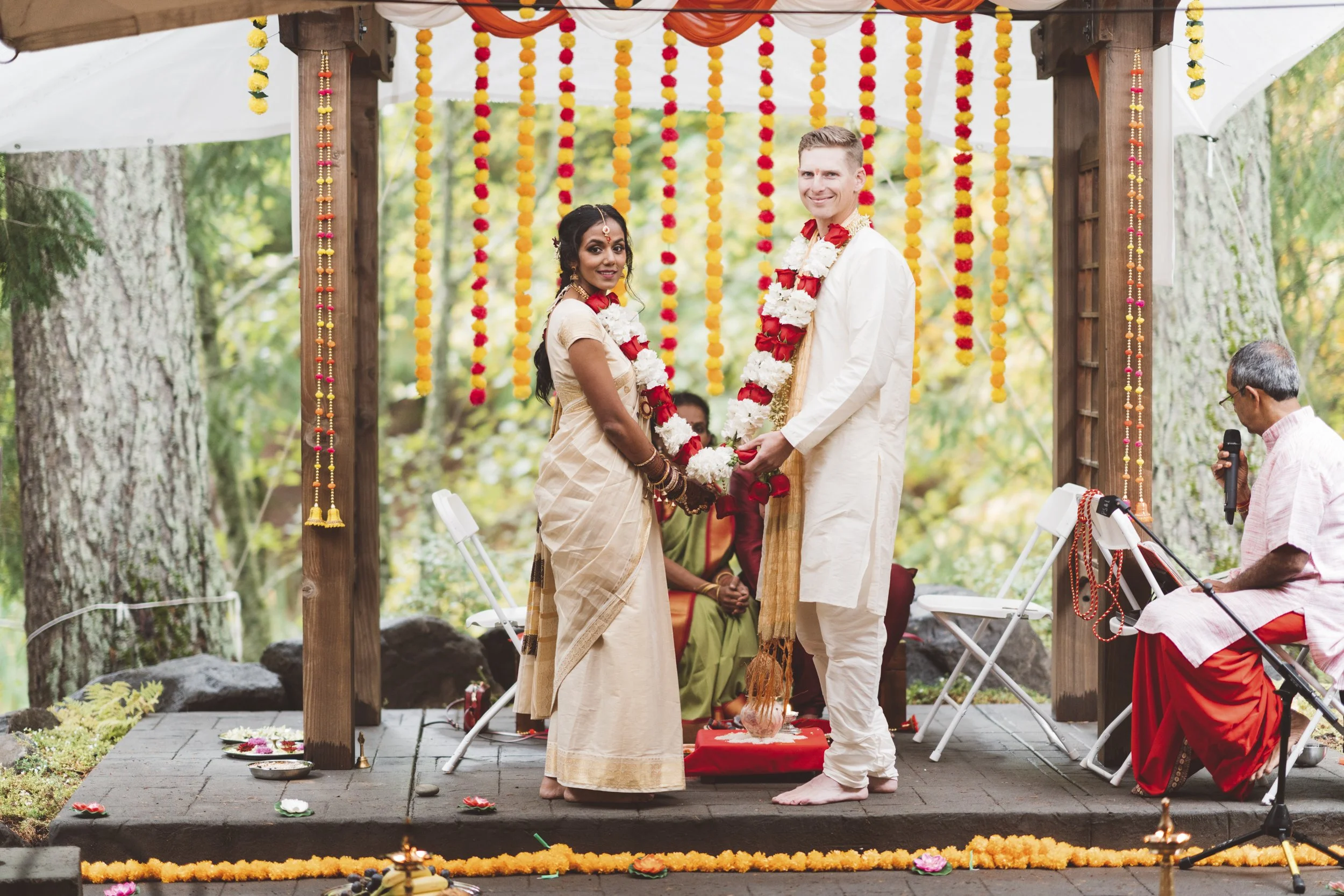 A couple dressed in traditional Indian attire holding hands during a wedding ceremony, standing on a decorated outdoor stage with marigold floral decorations hanging above.