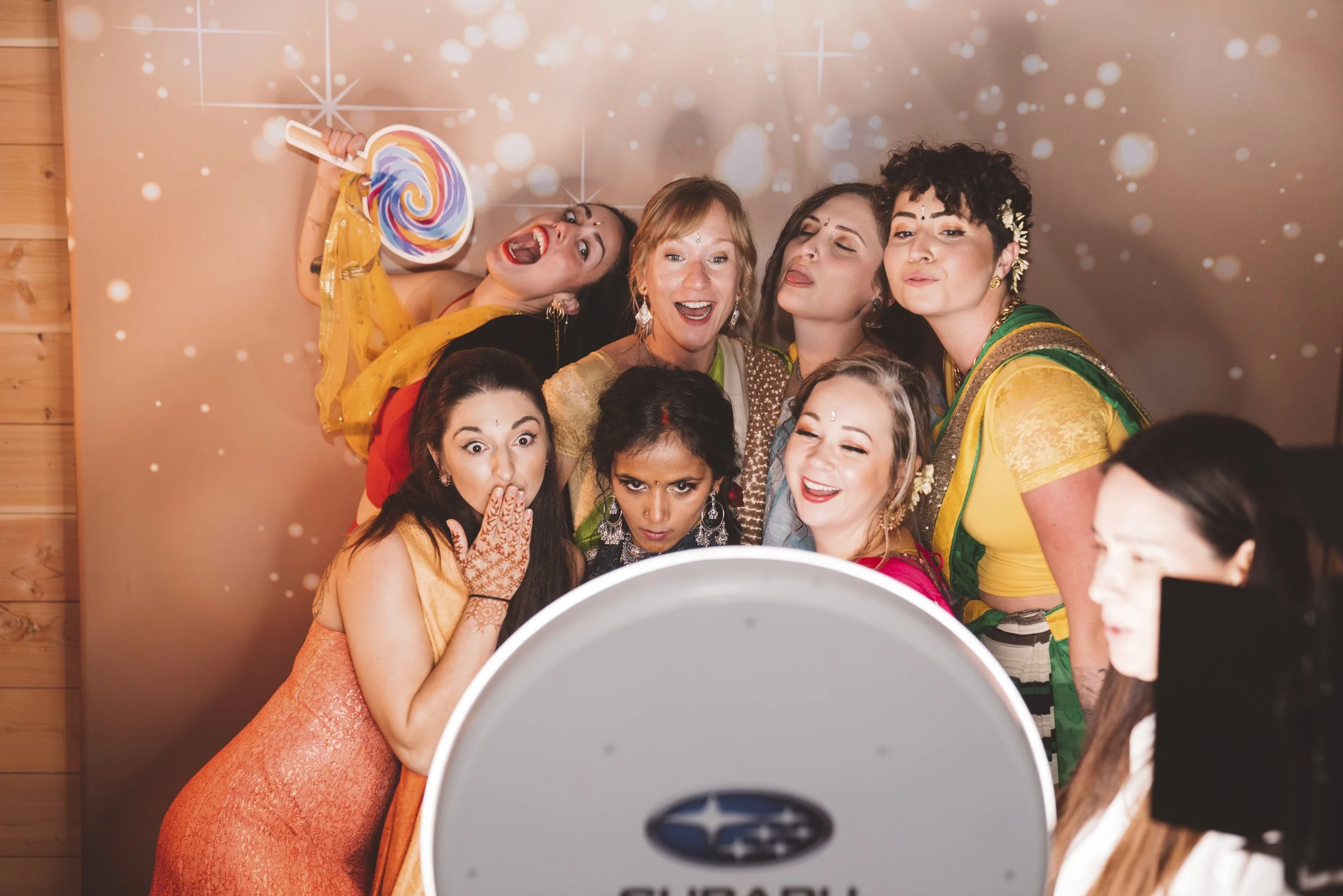 Group of women taking a selfie at a celebration, some making funny faces, in colorful traditional Indian attire, with a starry background and a camera lens visible in the foreground.