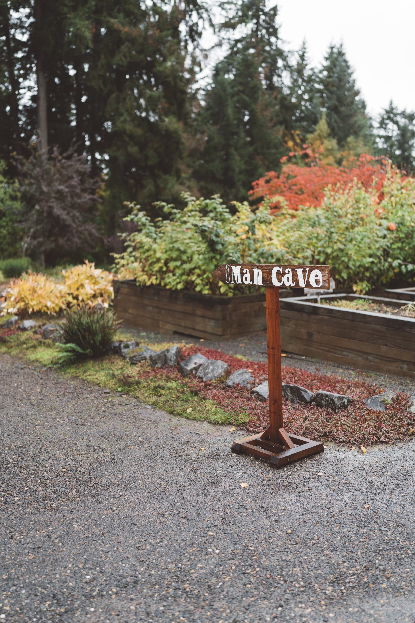 A wooden sign pointing to the Man Cave, placed on a gravel pathway, with colorful autumn bushes and trees in the background.
