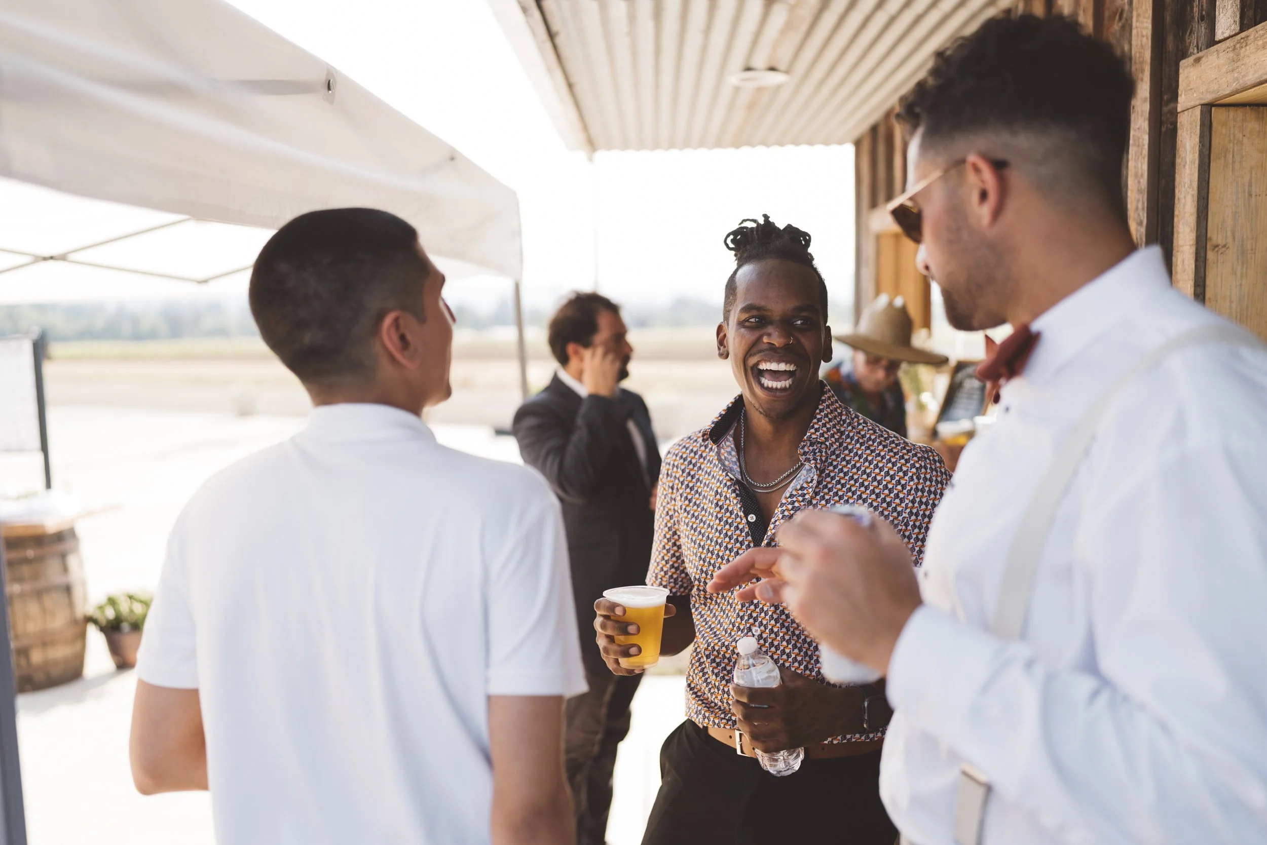 Group of people socializing outdoors, smiling and enjoying beverages, with a rustic wooden building in the background.