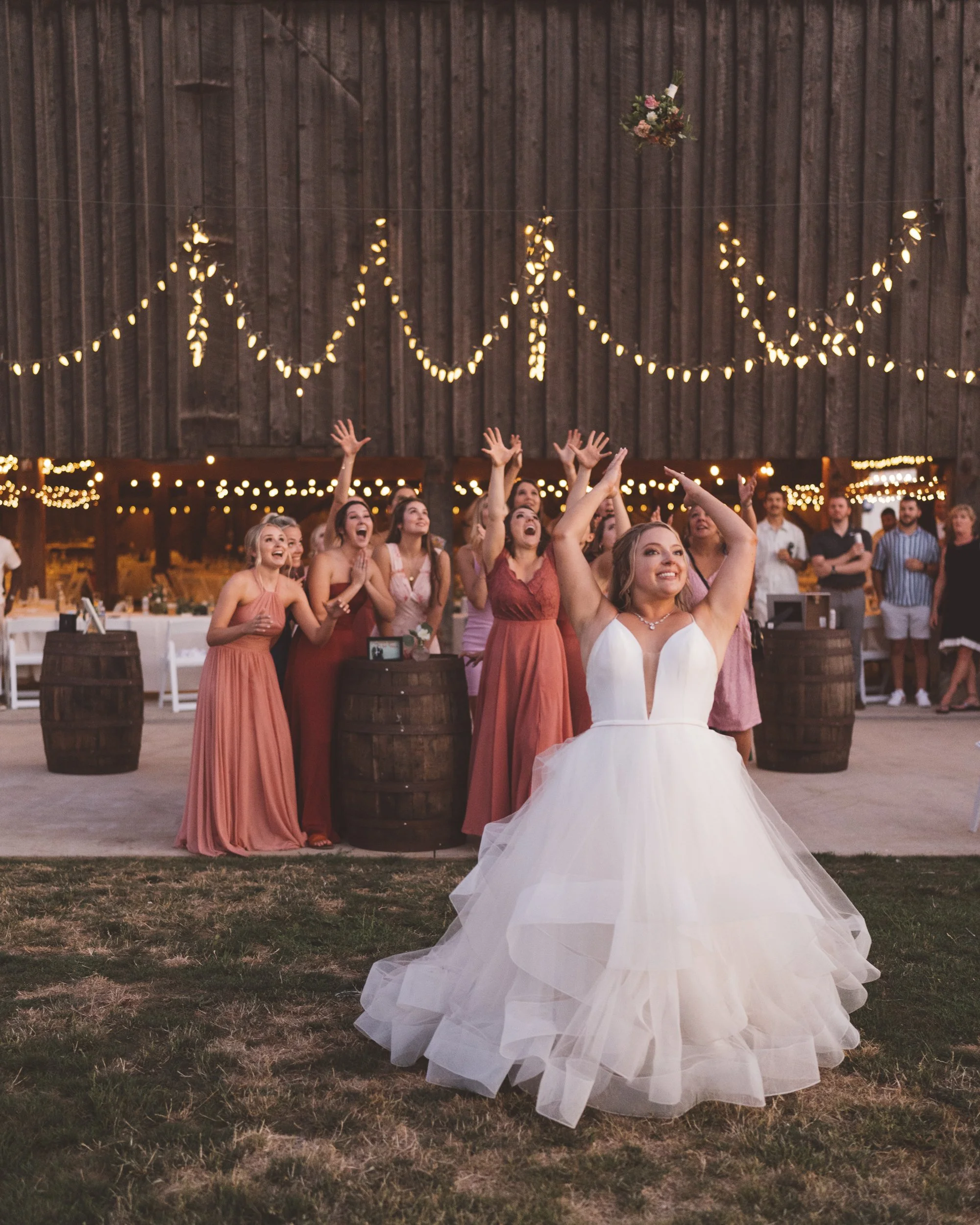 A bride in a white wedding gown tossing her bouquet to a group of women in peach and pink dresses at an outdoor wedding reception with string lights and a rustic barn backdrop.