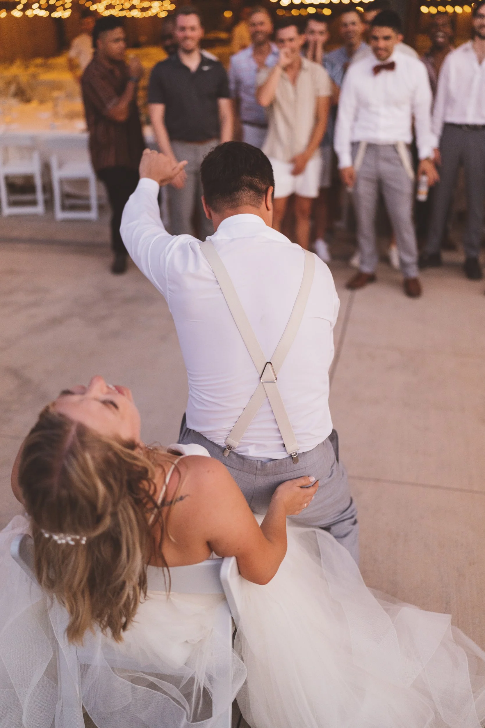 At a wedding reception, a groom in a white shirt and suspenders is doing the limbo as the bride, in a white dress, laughs and leans back in her chair. Guests stand in the background watching and smiling.