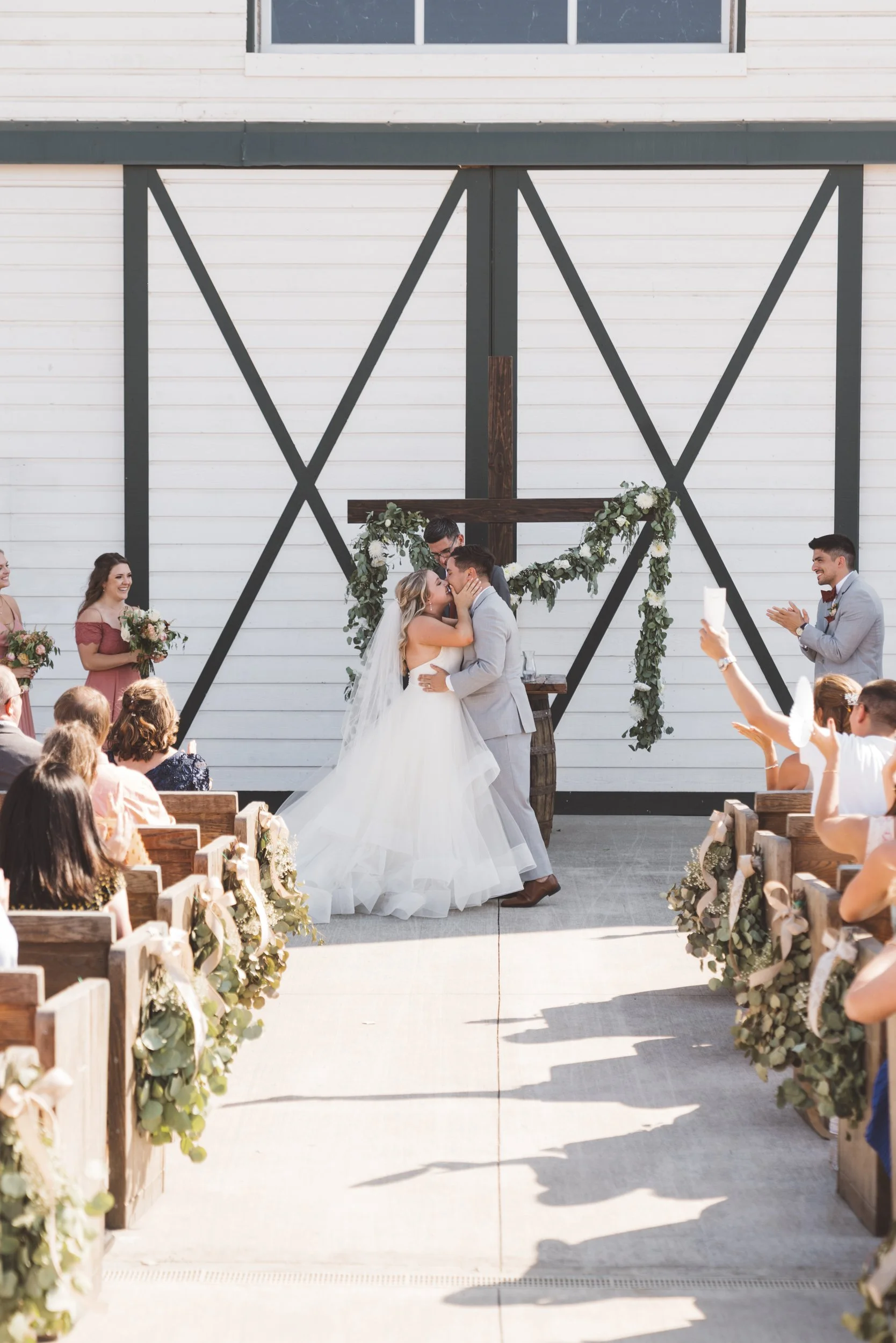 A wedding ceremony with a couple kissing in front of a cross decorated with greenery and flowers, surrounded by seated guests and bridesmaids, outside a barn with barn doors in the background.