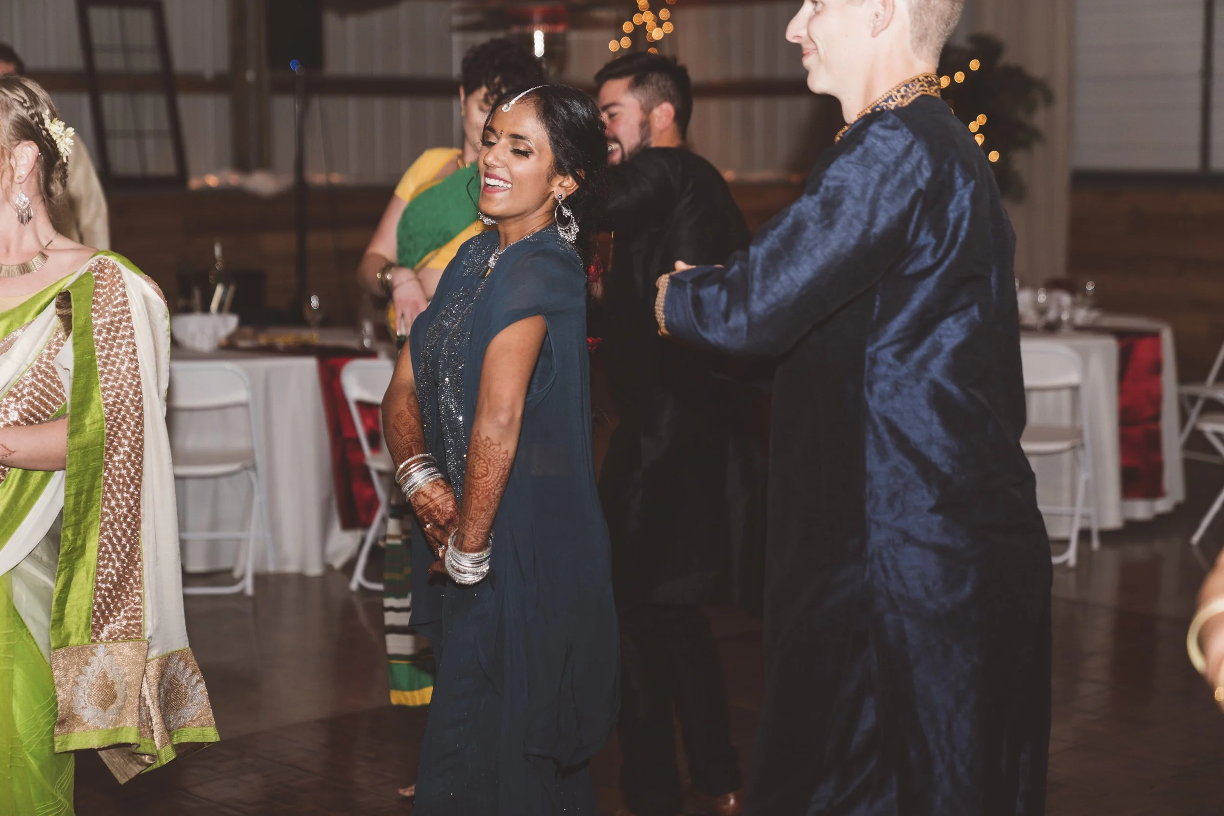 Group of people dancing and celebrating at an indoor event, with women wearing traditional Indian attire and henna on their hands.