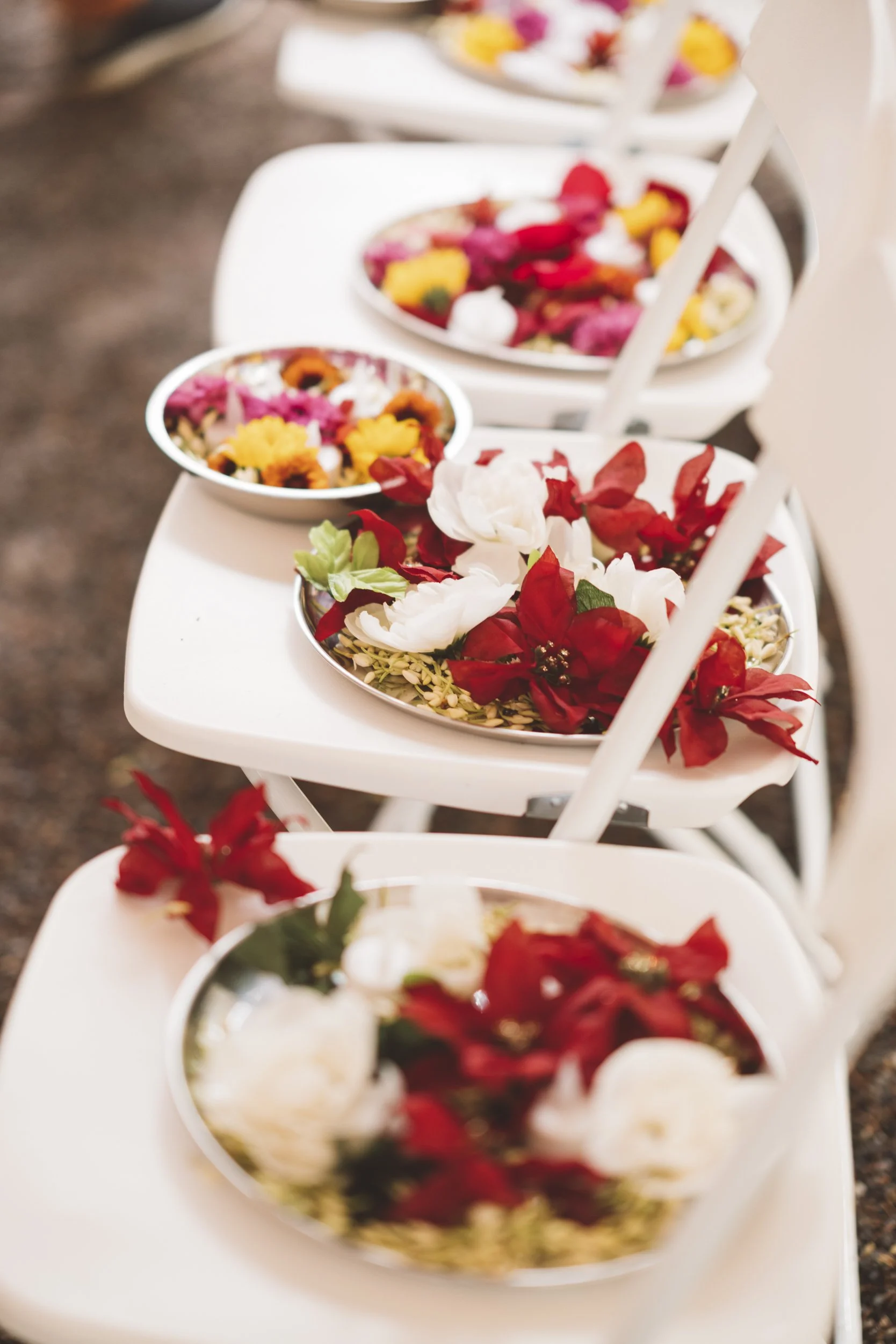 White chairs with small bowls of colorful flower petals on a table, focusing on the front bowl with red, white, and green flowers.
