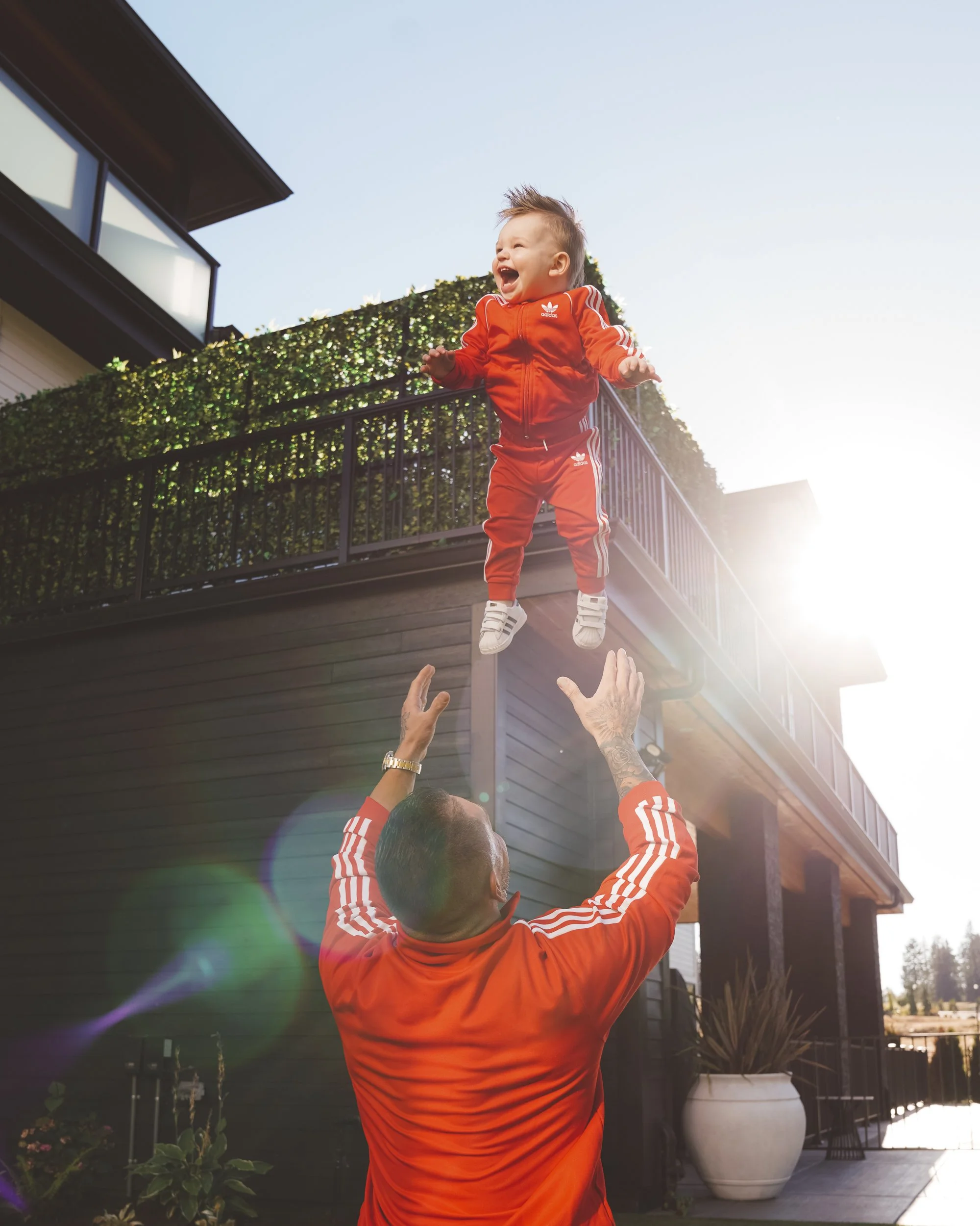 A young child dressed in a red Adidas tracksuit being thrown into the air by an adult, likely a parent, on a sunny day outside a modern house with a balcony and plants.