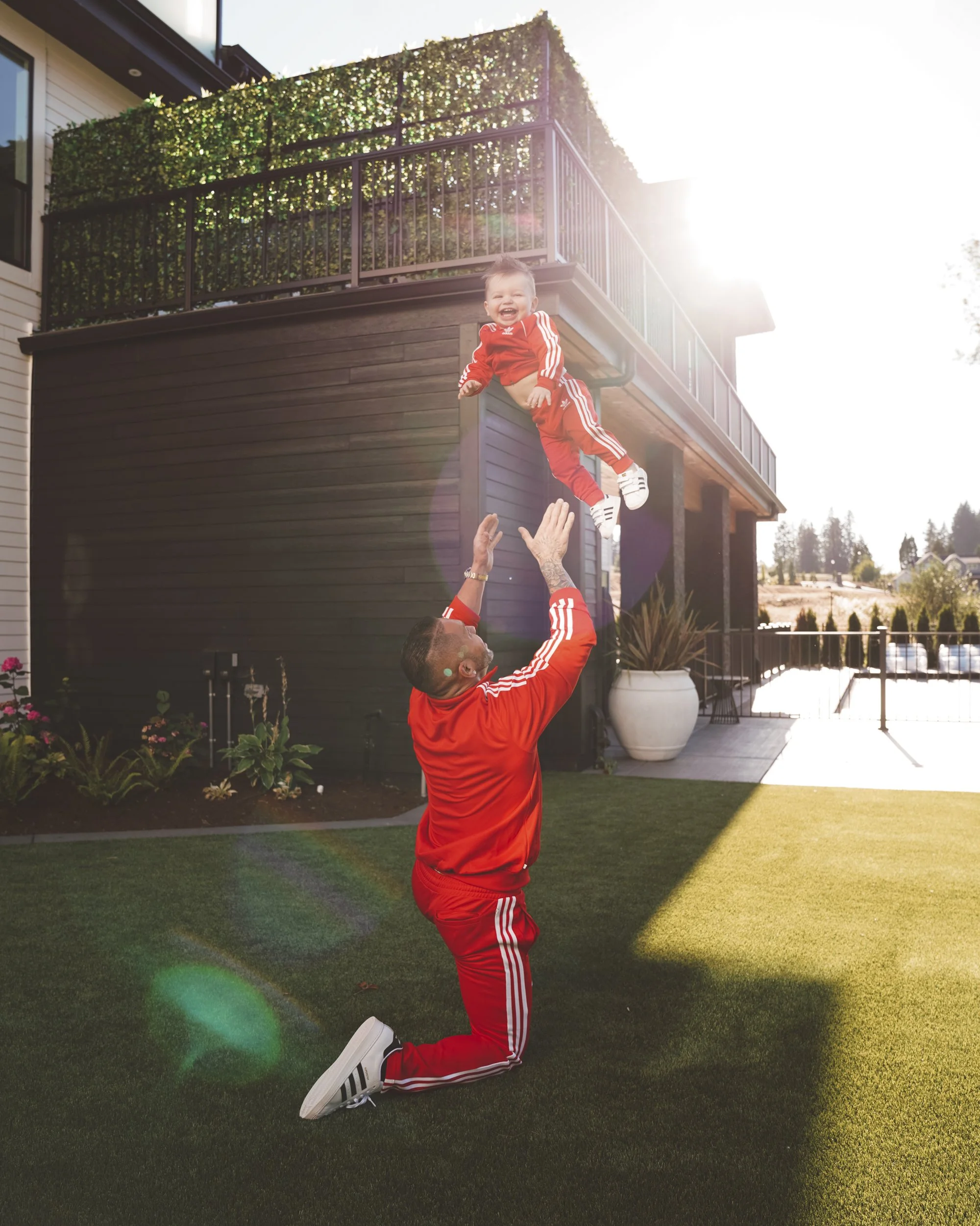 A man in a red tracksuit is kneeling outdoors on a green lawn, tossing a young boy dressed in red into the air. The boy is smiling widely with sunlight in the background and a house with a deck and plants behind them.