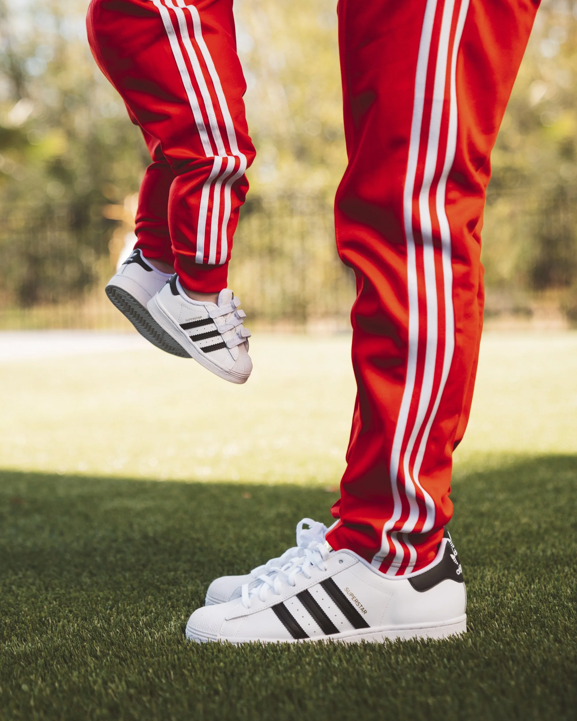 A person wearing white Adidas sneakers and red track pants with white stripes, holding a child's foot, outdoors on grass with trees in the background.