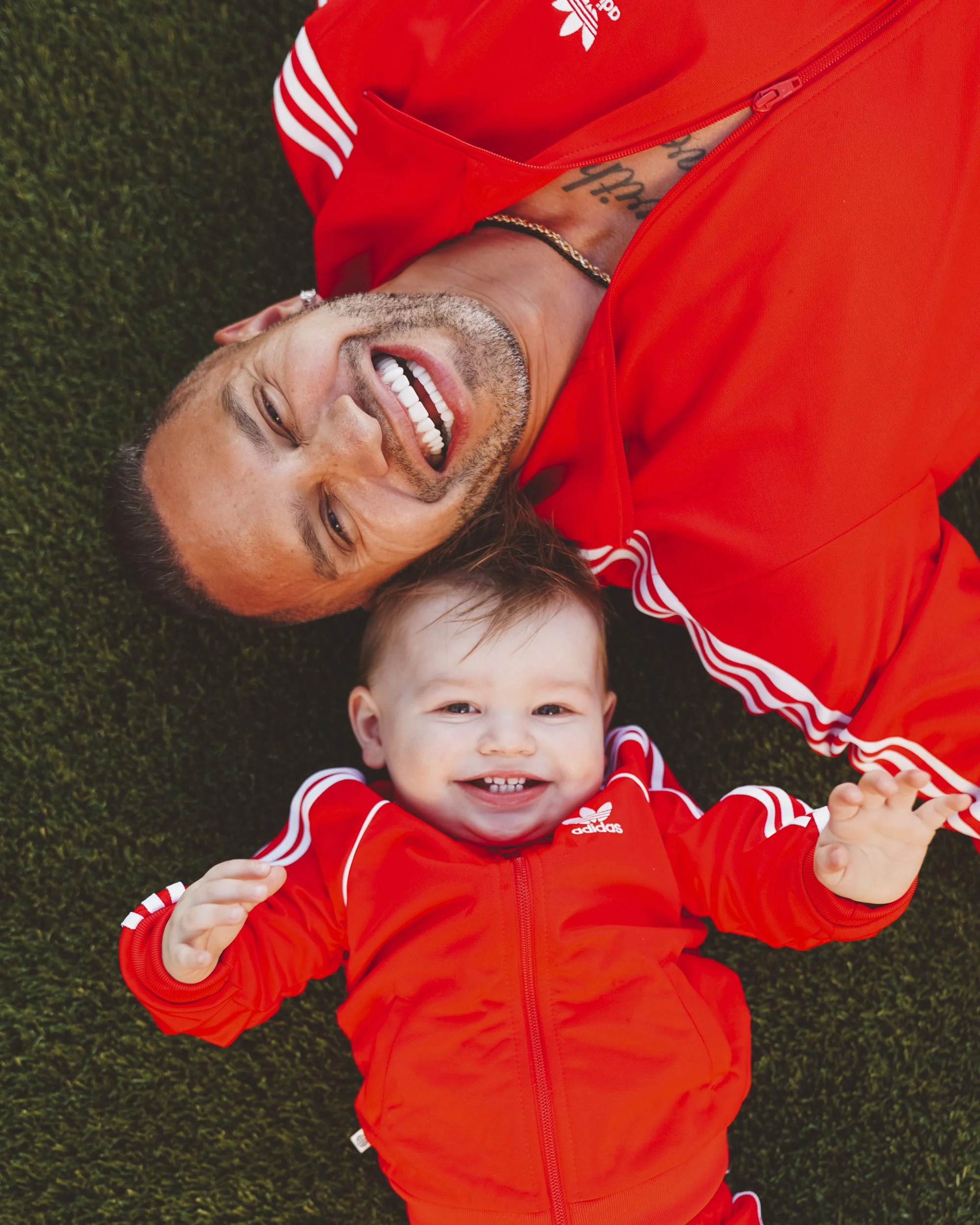 A man and a smiling baby lying on grass, both wearing matching red Adidas tracksuits.