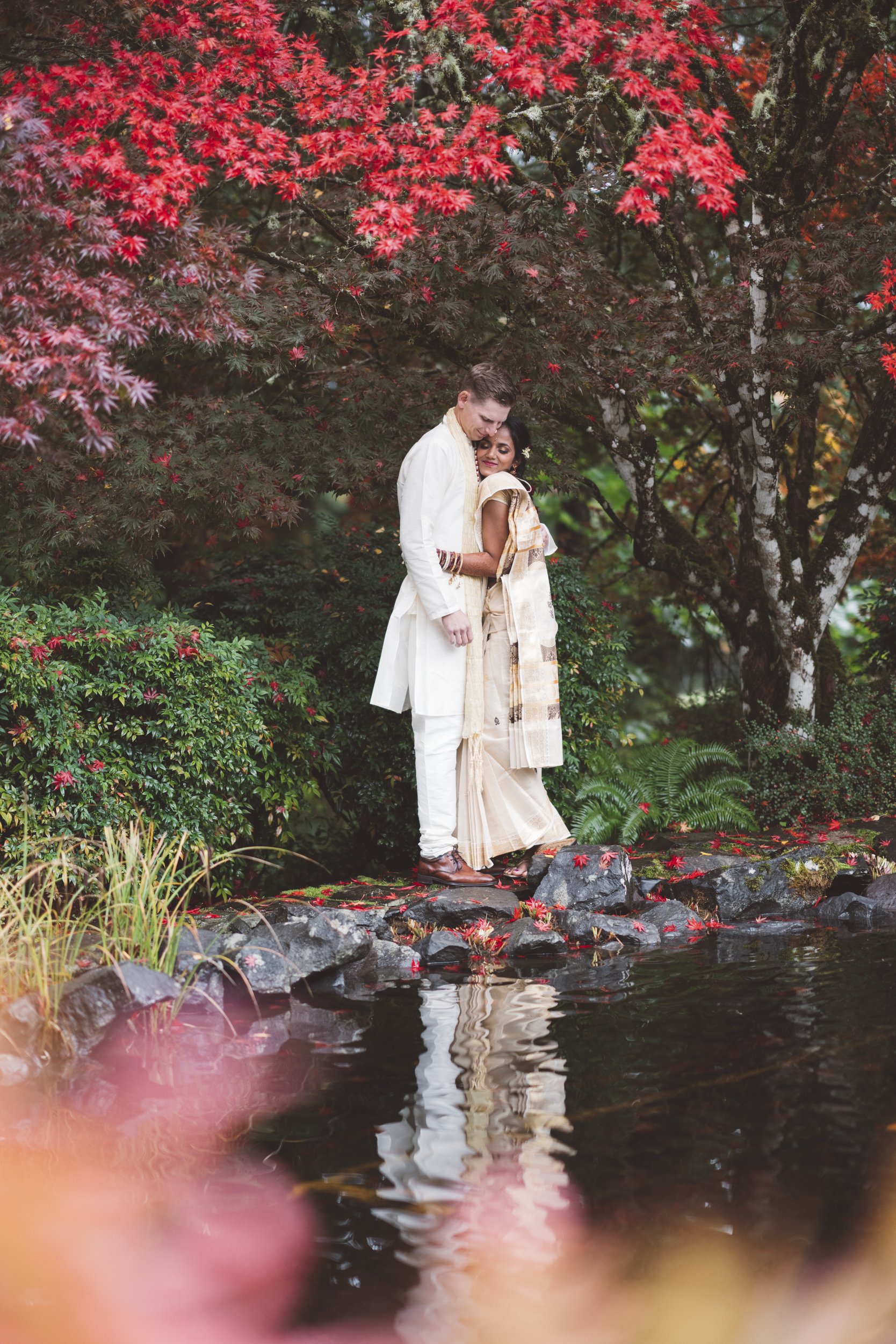 A couple dressed in traditional Indian attire standing on rocks by a pond surrounded by lush greenery and red autumn leaves, embracing each other lovingly.