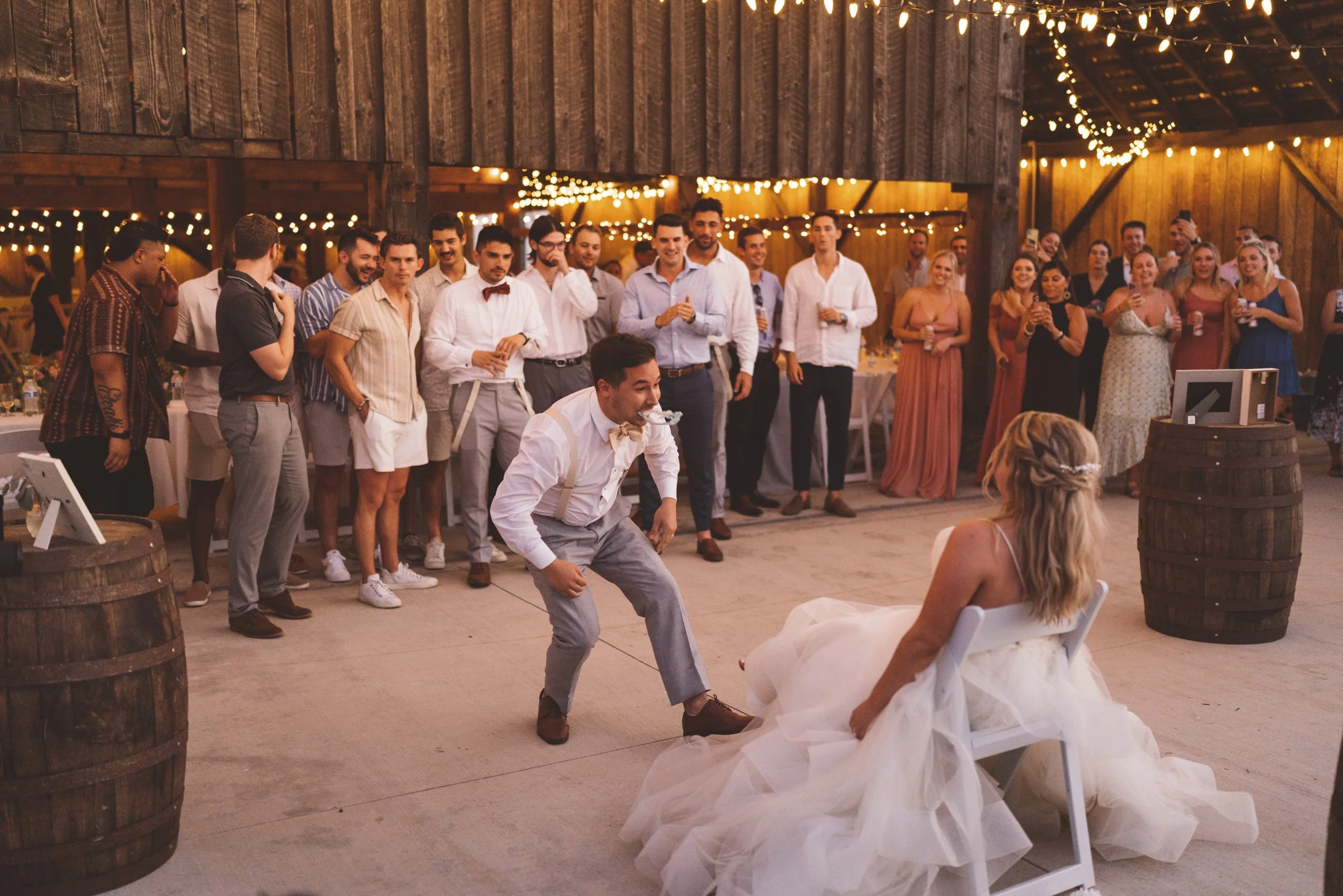 At a wedding reception, a groom in suspenders and dress shoes is about to kiss the bride, who is sitting on a chair wearing a white wedding dress. The guests, dressed in semi-formal attire, stand in the background under string lights in a rustic barn