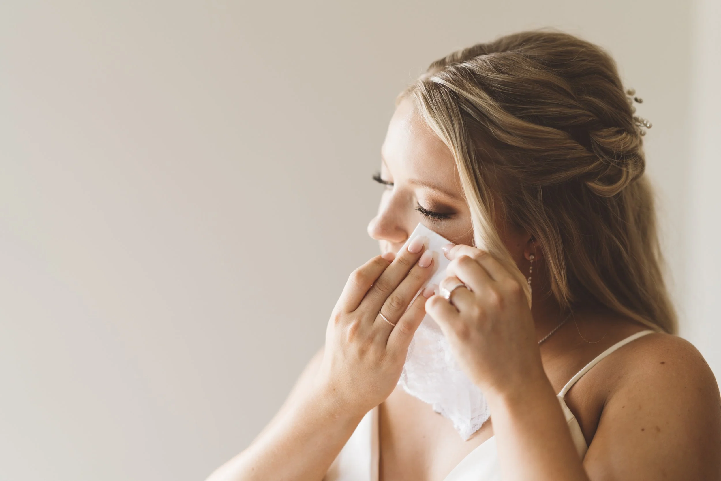 A woman with styled blonde hair and earrings, wearing a white sleeveless top, is using a white tissue or cloth to wipe her face or nose against a plain light background.