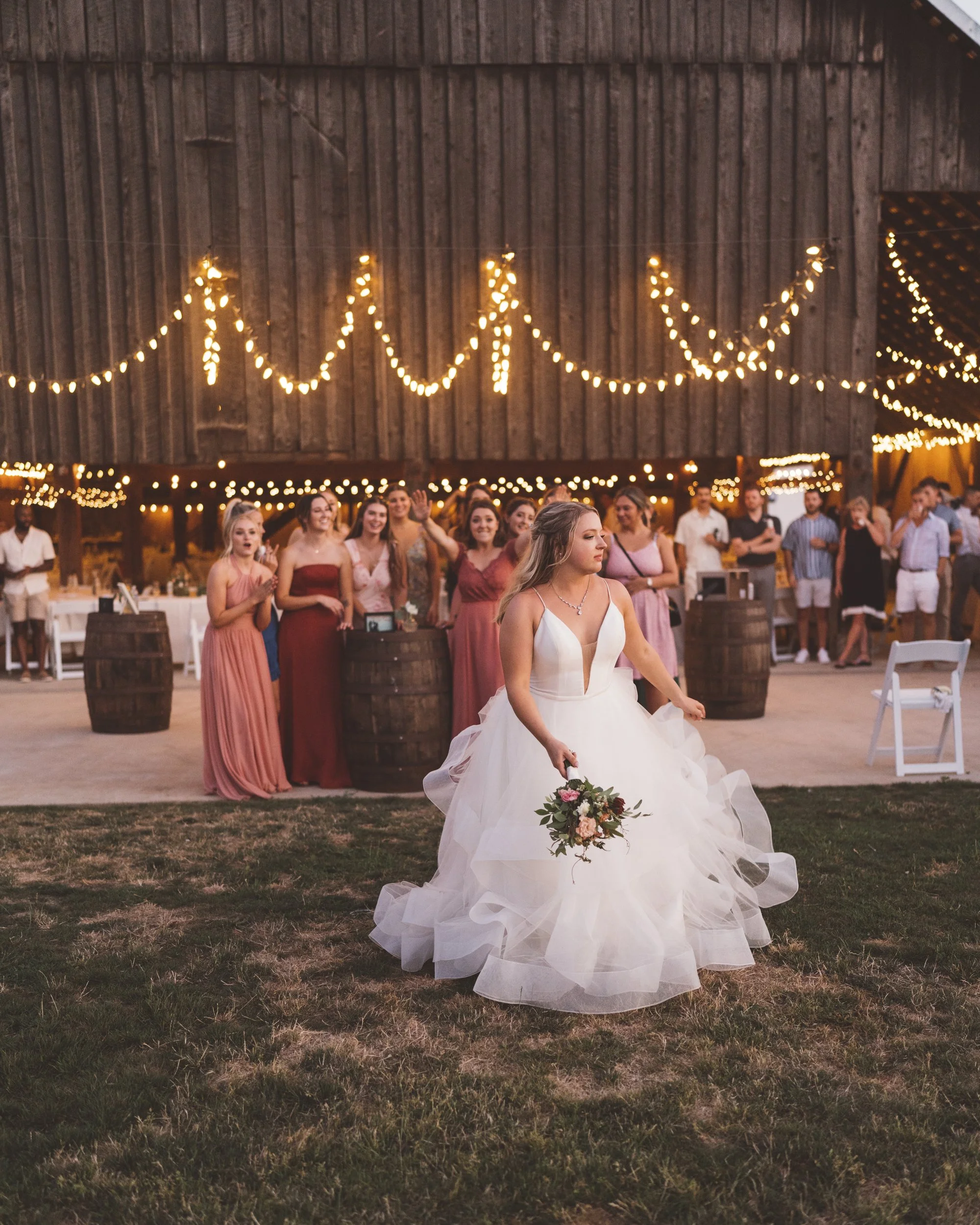 Bride in wedding gown holding bouquet standing on grass, with bridesmaids and guests in background under string lights at a barn wedding reception.