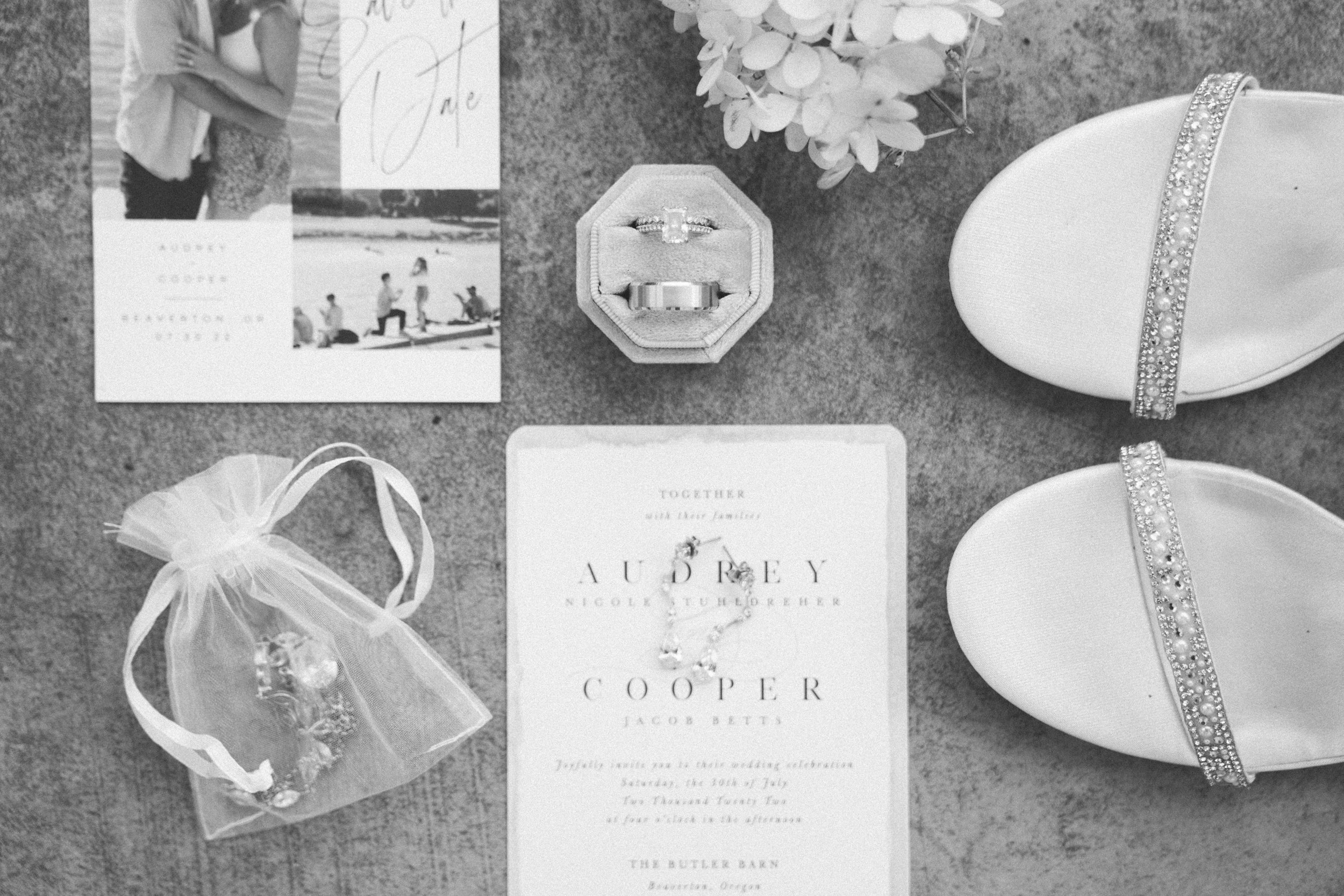 Wedding invitation, engagement ring in box, wedding bands, small gift bag, bouquet, and white shoes with embellished straps