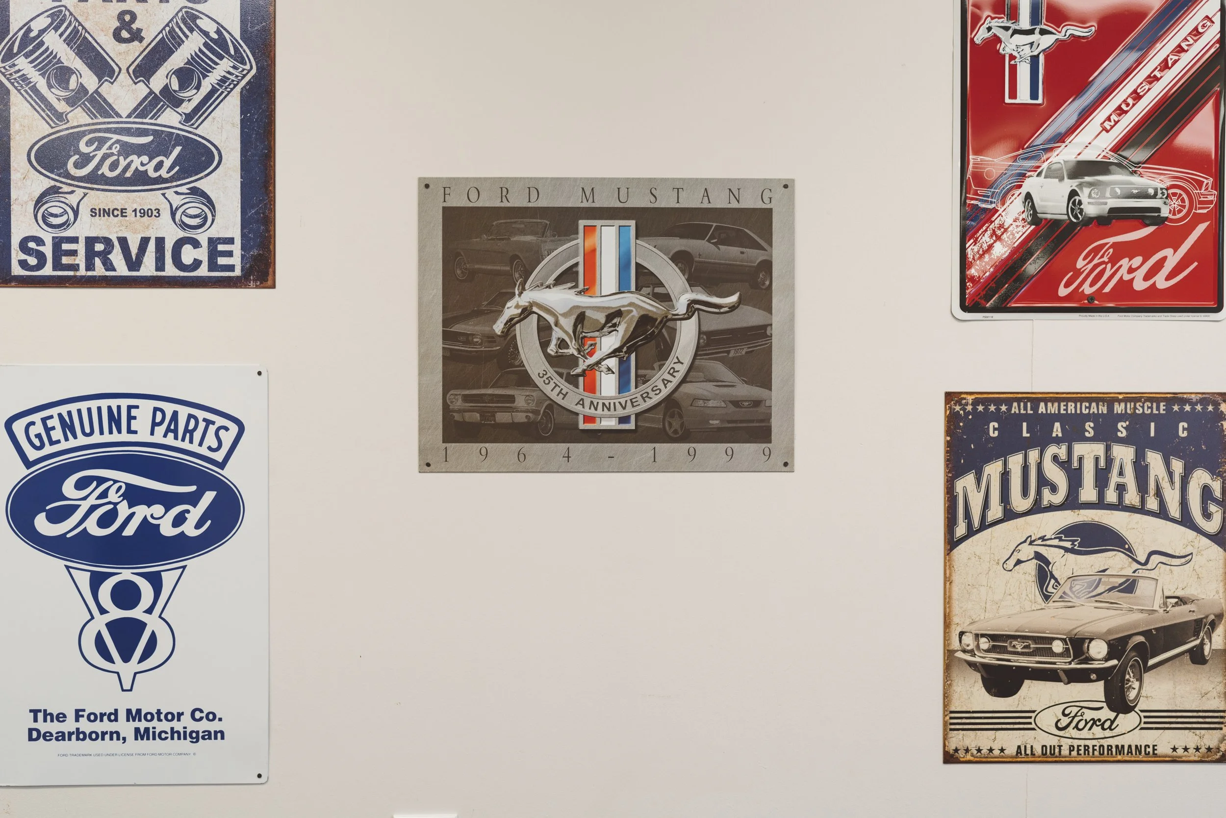 A white wall with five vintage Ford Mustang posters. The top left poster is blue and white, advertising Ford service since 1903. Center is a beige poster celebrating the 35th anniversary of the Ford Mustang, featuring a silver Mustang logo with image