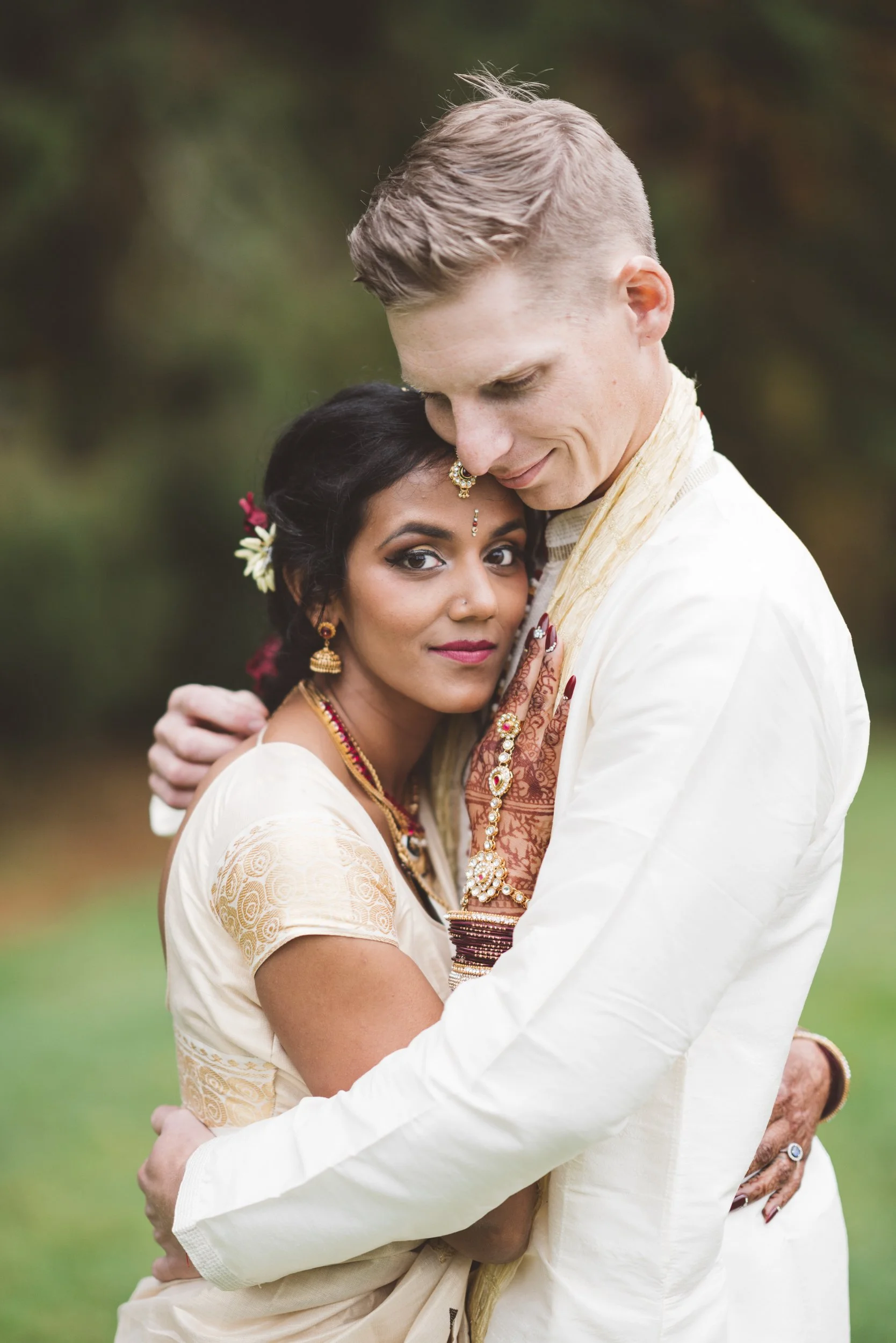 A couple hugging outdoors with the woman looking at the camera and the man with his head bowed, wearing traditional Indian attire.