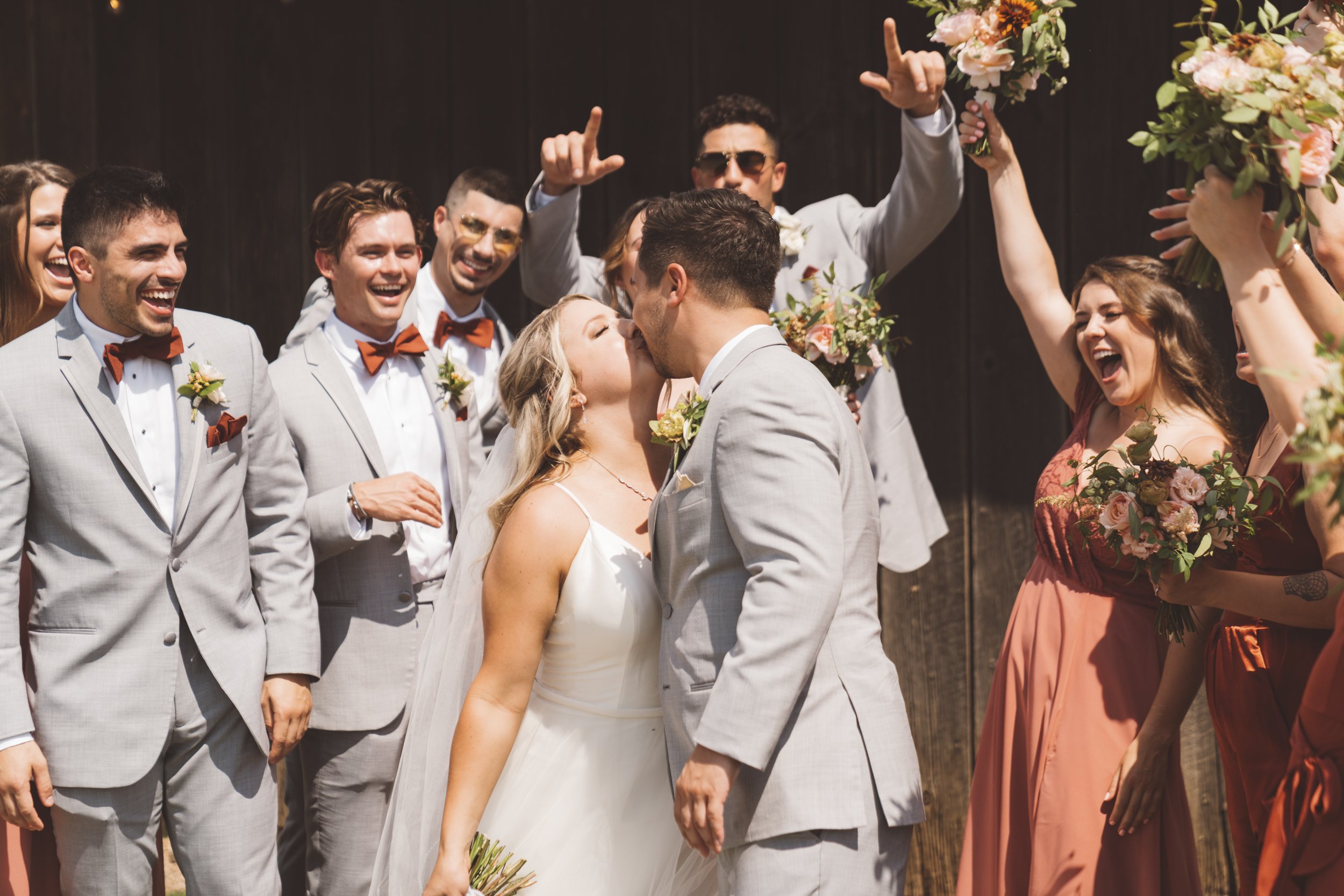 A wedding couple sharing a kiss surrounded by laughing friends and bridesmaids holding bouquets.