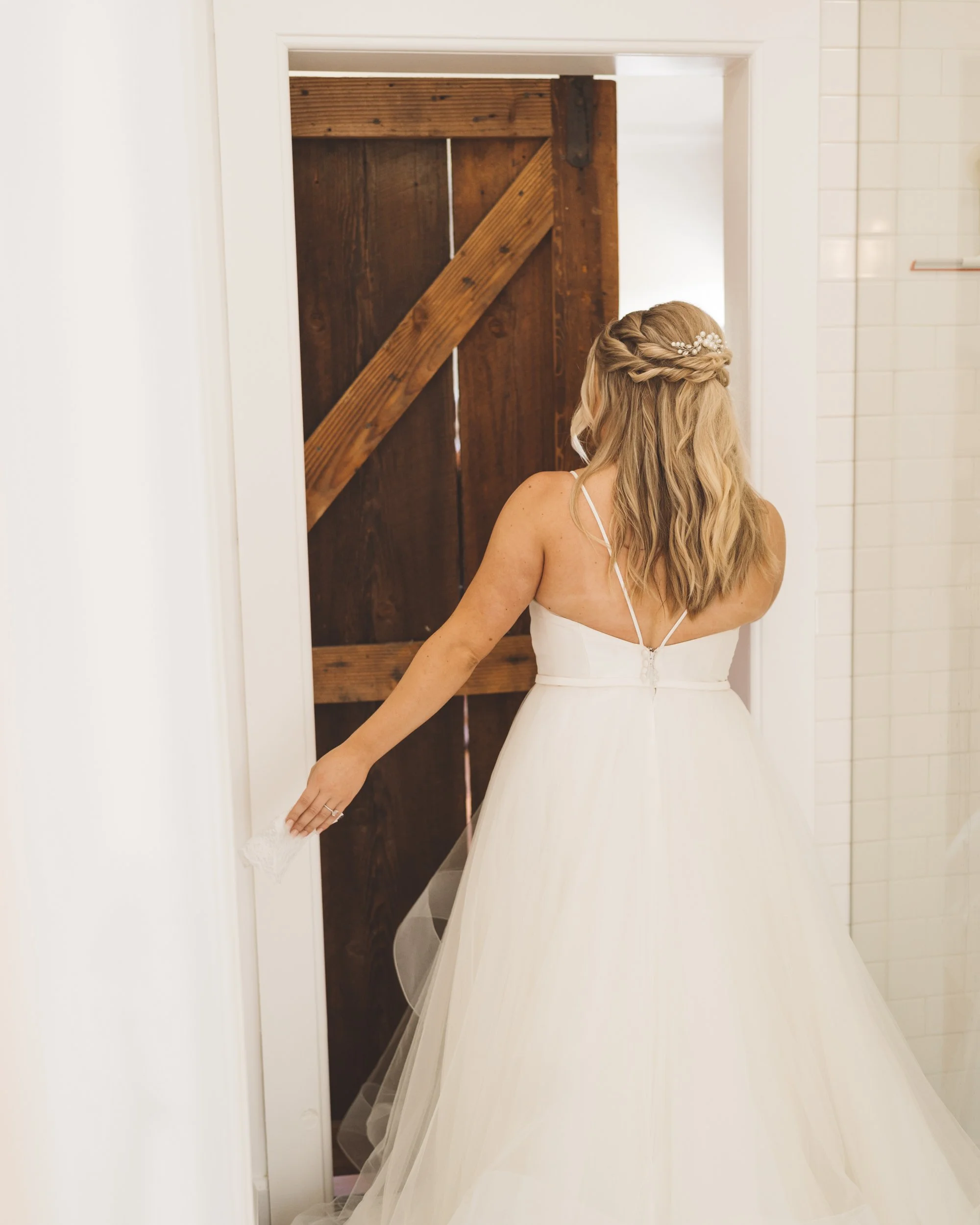 Bride in a white wedding dress with spaghetti straps opening a wooden barn door.