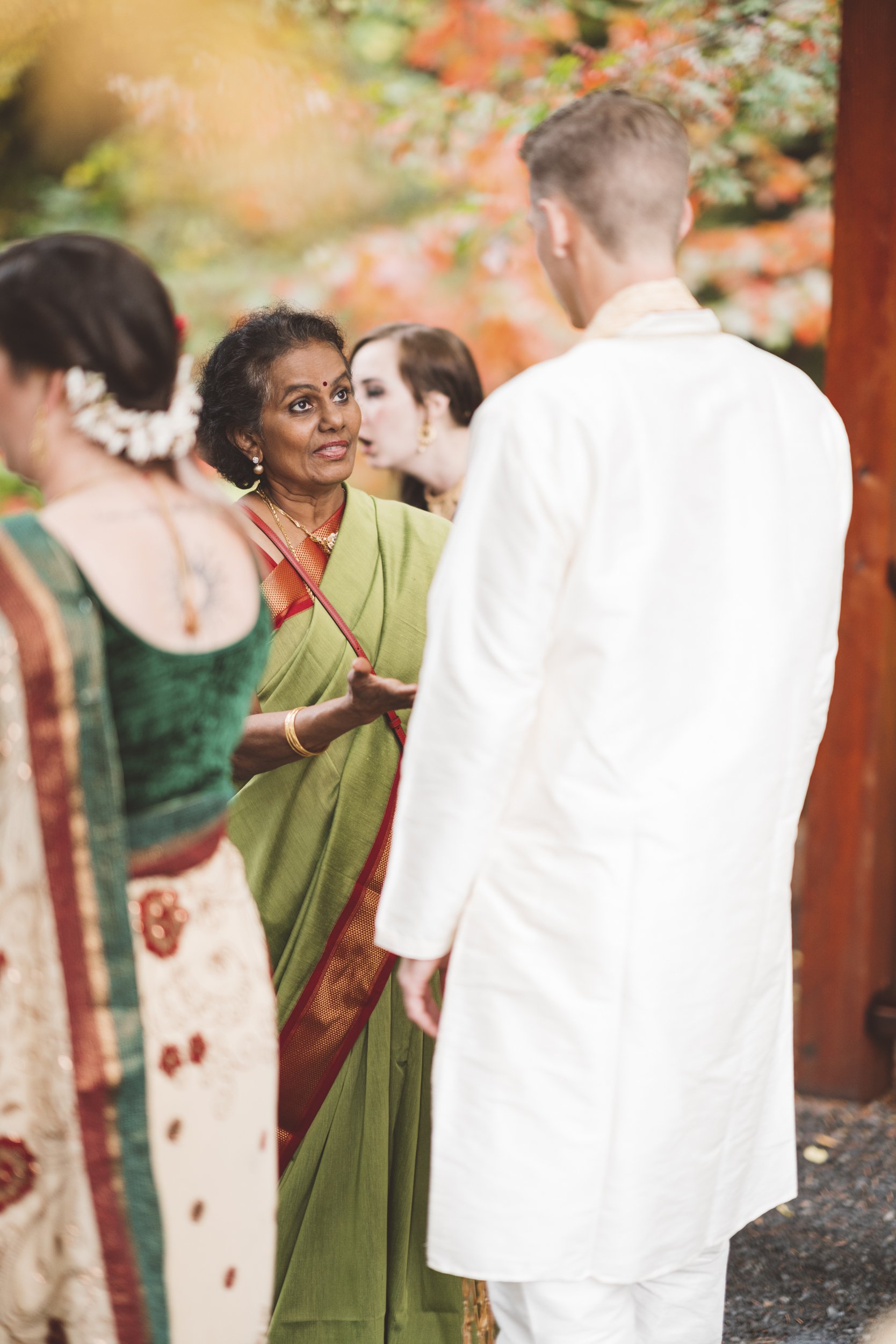 An Indian woman in a green saree talking to a man in a white traditional outfit at an outdoor event with autumn-colored trees in the background.