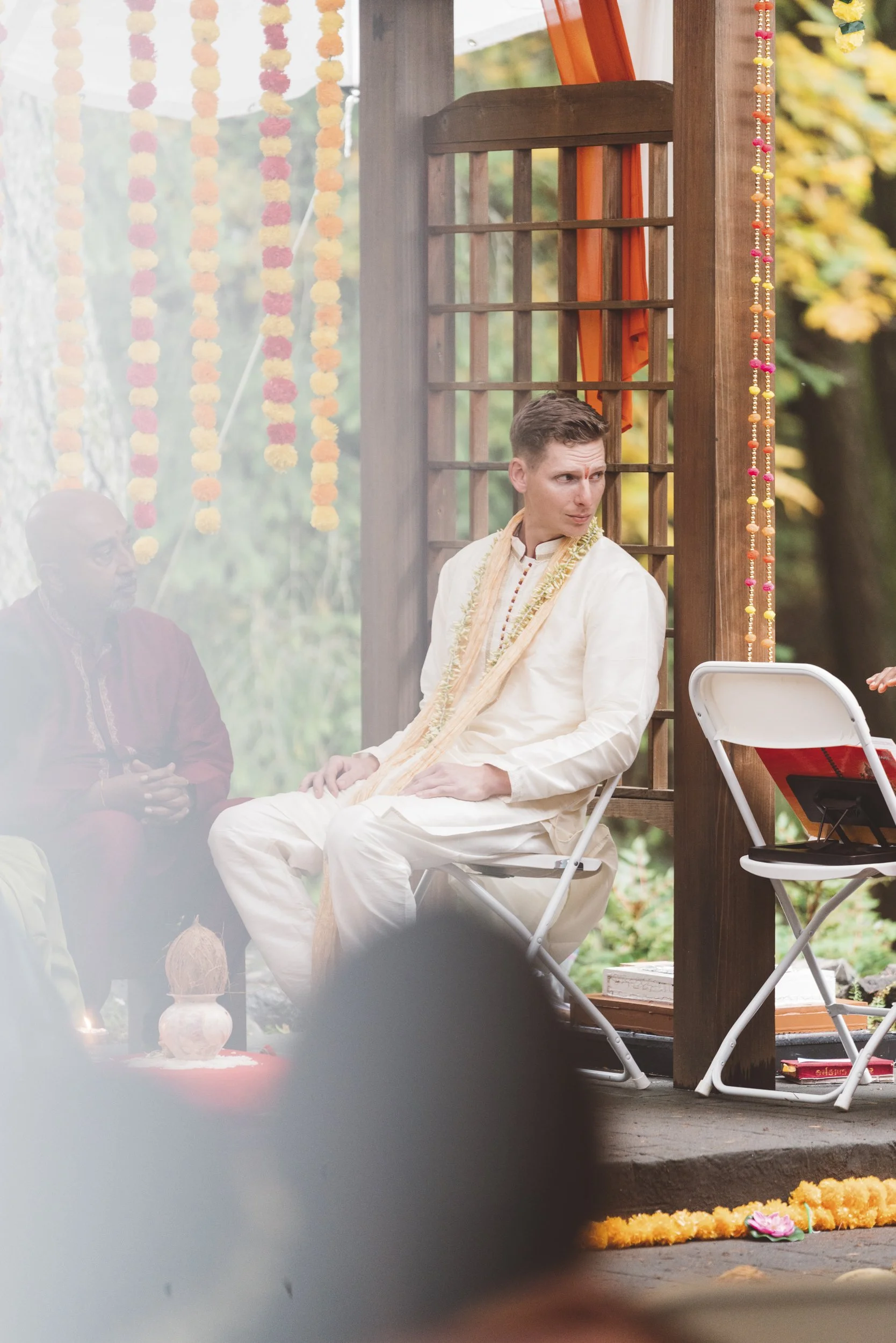 Man dressed in traditional Indian attire sitting on a white foldable chair during a cultural event, with a woman in the background and colorful decorations around him.