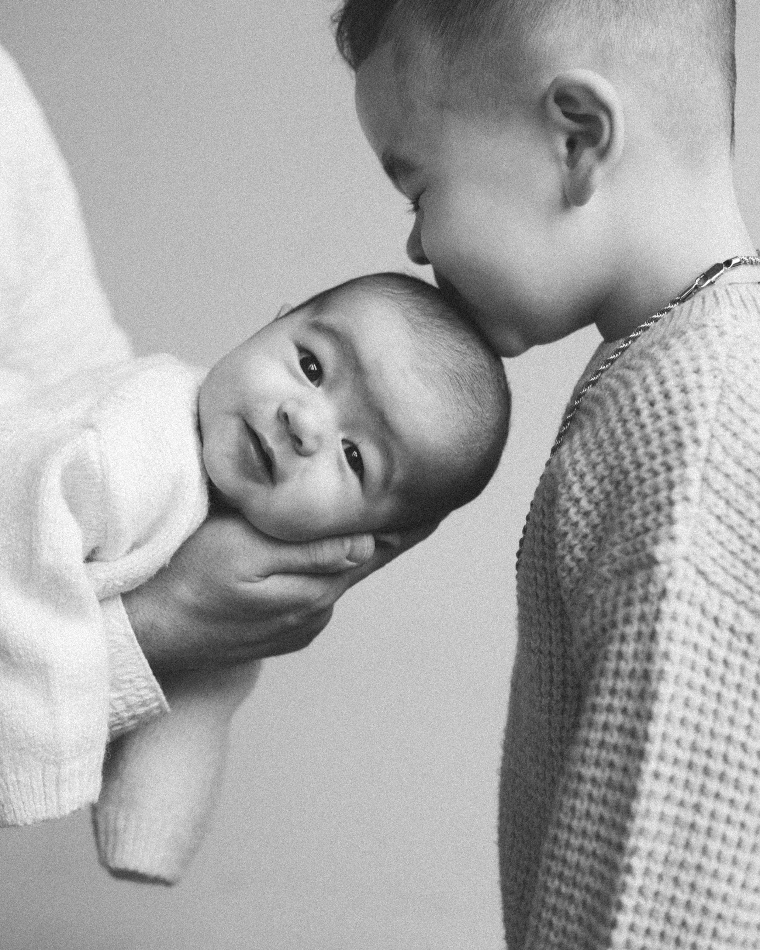 A young boy kisses a baby on the head while holding the baby, with a neutral background.
