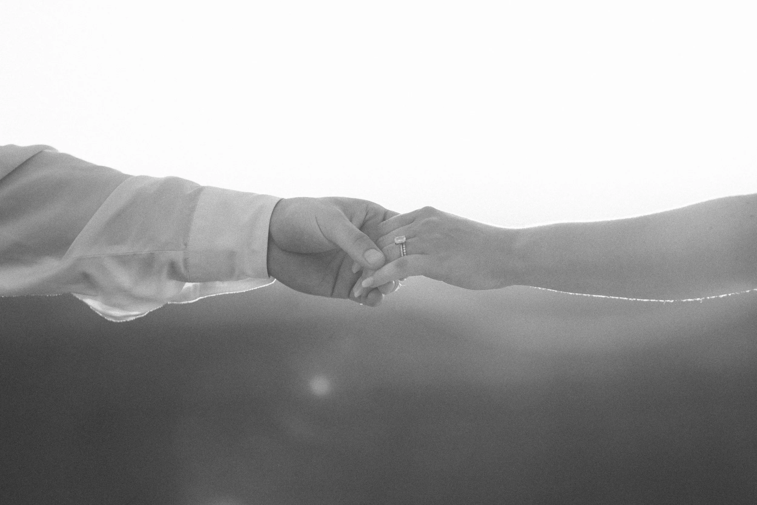 A close-up of a bride and groom holding hands, with the bride wearing rings, against a plain background.